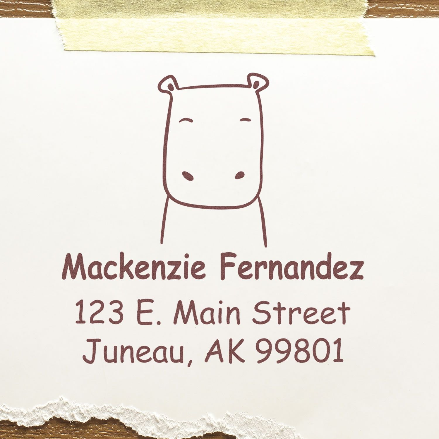Portable Pre-Inked Hopeful Hippo Made To Order Mailing Stamp on paper with a hippo illustration. Customized Address Stamp reads Mackenzie Fernandez, 123 E. Main Street, Juneau, AK 99801 in brown ink, taped to a wooden surface.