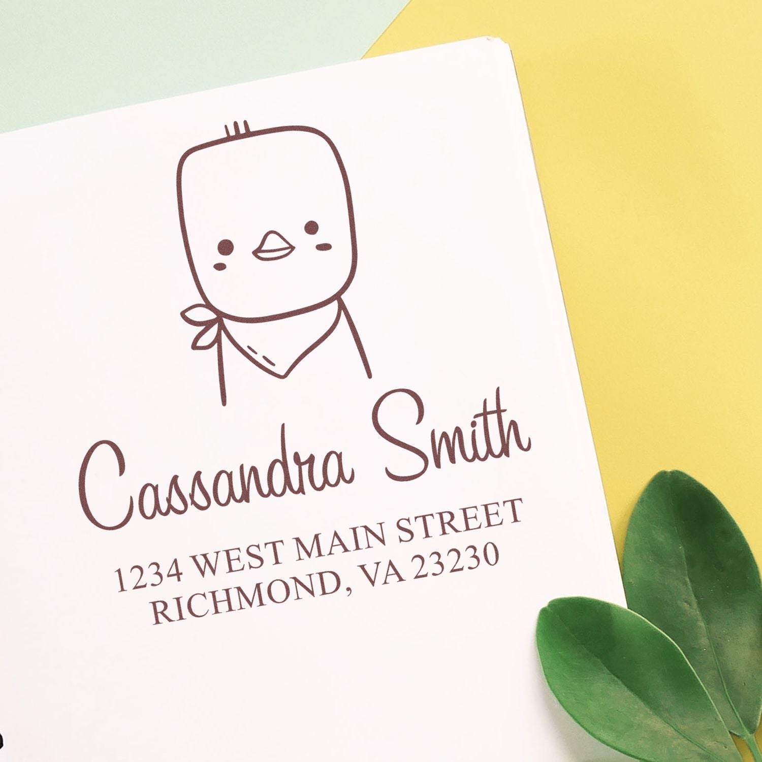 Image of a Portable Pre-Inked Cute Chick Made To Order Address Stamper imprint on paper. The design features a cartoon chick above the name "Cassandra Smith" and an address. This Personalizable Mailing Stamper is shown with green leaves and a yellow background.