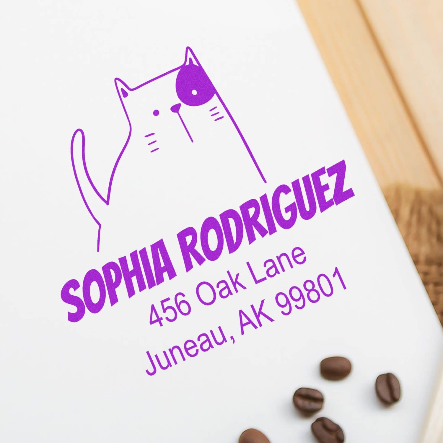 Wood handle fluffy feline cartoon animal custom envelope stamper in use, featuring a purple cat design. Customized return mail stamp displays name and address on a white envelope, surrounded by coffee beans on a wooden surface.