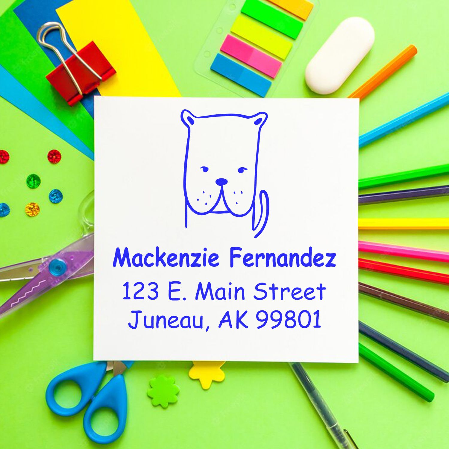 Self-Inking Cartoon Animal Darling Dog Personalized Envelope Stamper displayed on a colorful desk with stationery. Custom Return Mail Stamp shows a cute dog illustration and personalized address for Mackenzie Fernandez, 123 E. Main Street, Juneau, AK 99801.