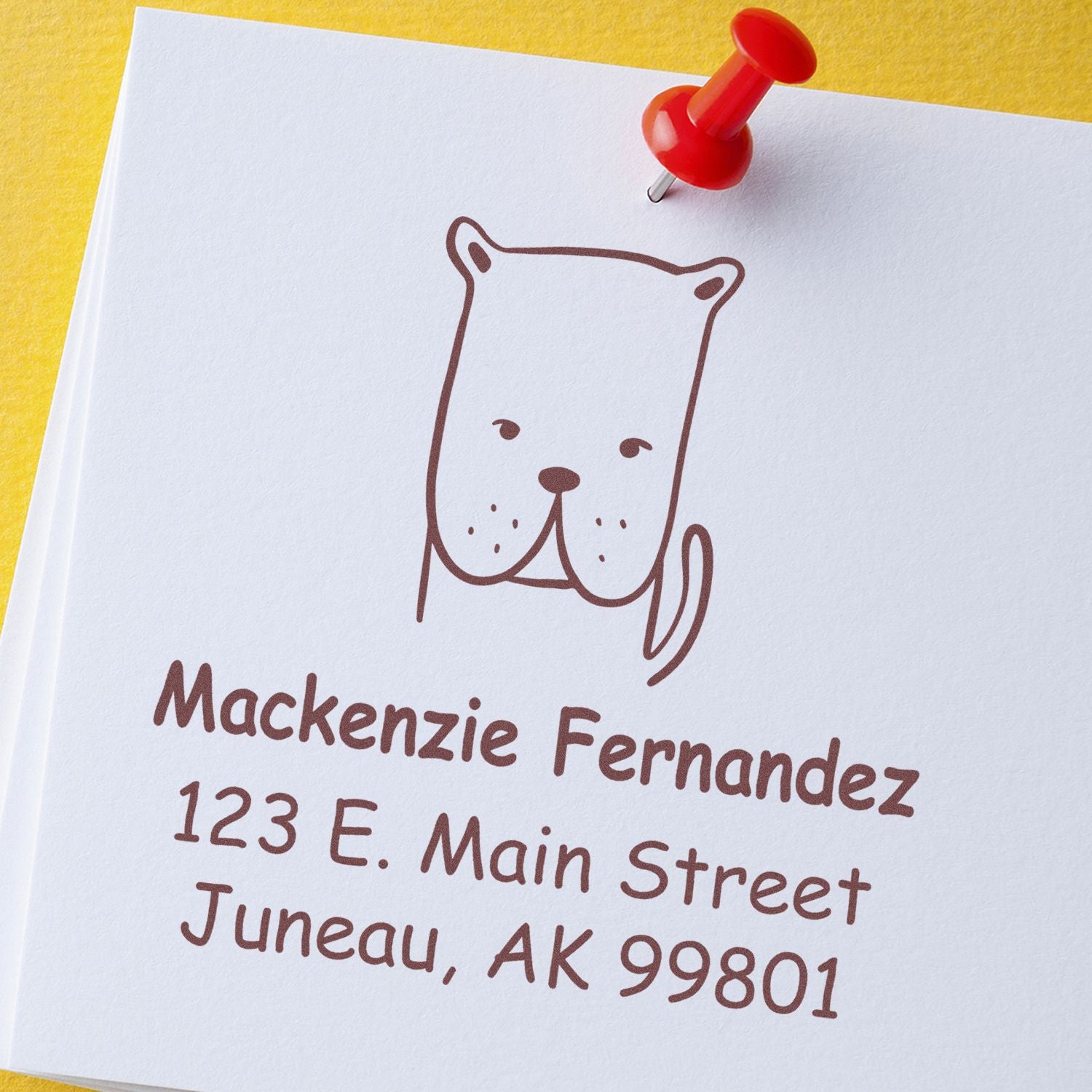 Wood handle darling dog cartoon animal custom address stamp on white paper with a red pushpin. Personalizable return mail stamp featuring a cute dog illustration and address details for Mackenzie Fernandez, 123 E. Main Street, Juneau, AK 99801.