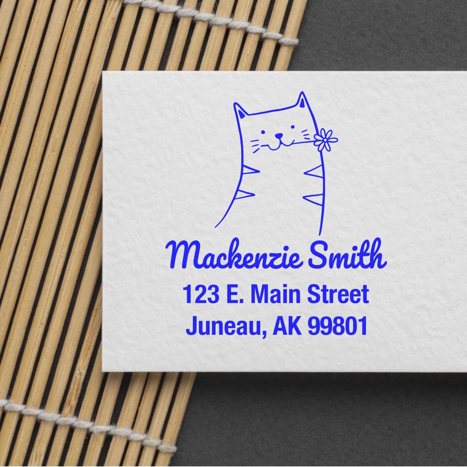 Wood handle happy cat cartoon animal custom address stamper on a customizable home stamp. The stamp features a smiling cat holding a flower, with the name "Mackenzie Smith" and an address in blue ink on a white card, placed on a bamboo mat.