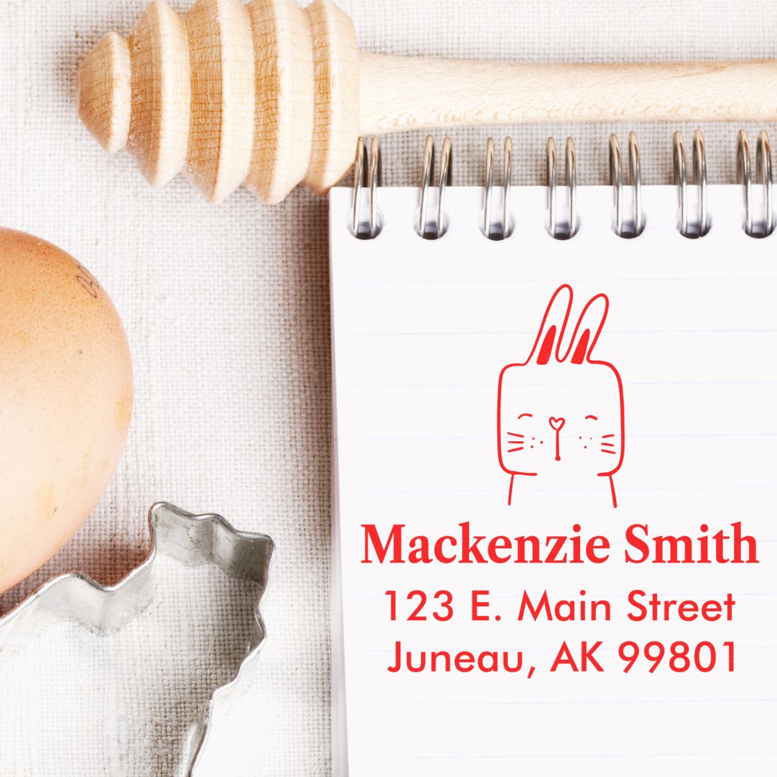 Fuzzy Rabbit Pre-Inked Handcrafted Address Stamp on a notepad with "Mackenzie Smith, 123 E. Main Street, Juneau, AK 99801" in red ink. Customizable Home Stamper design features a cute rabbit illustration. Nearby are a honey dipper, egg, and cookie cutter.