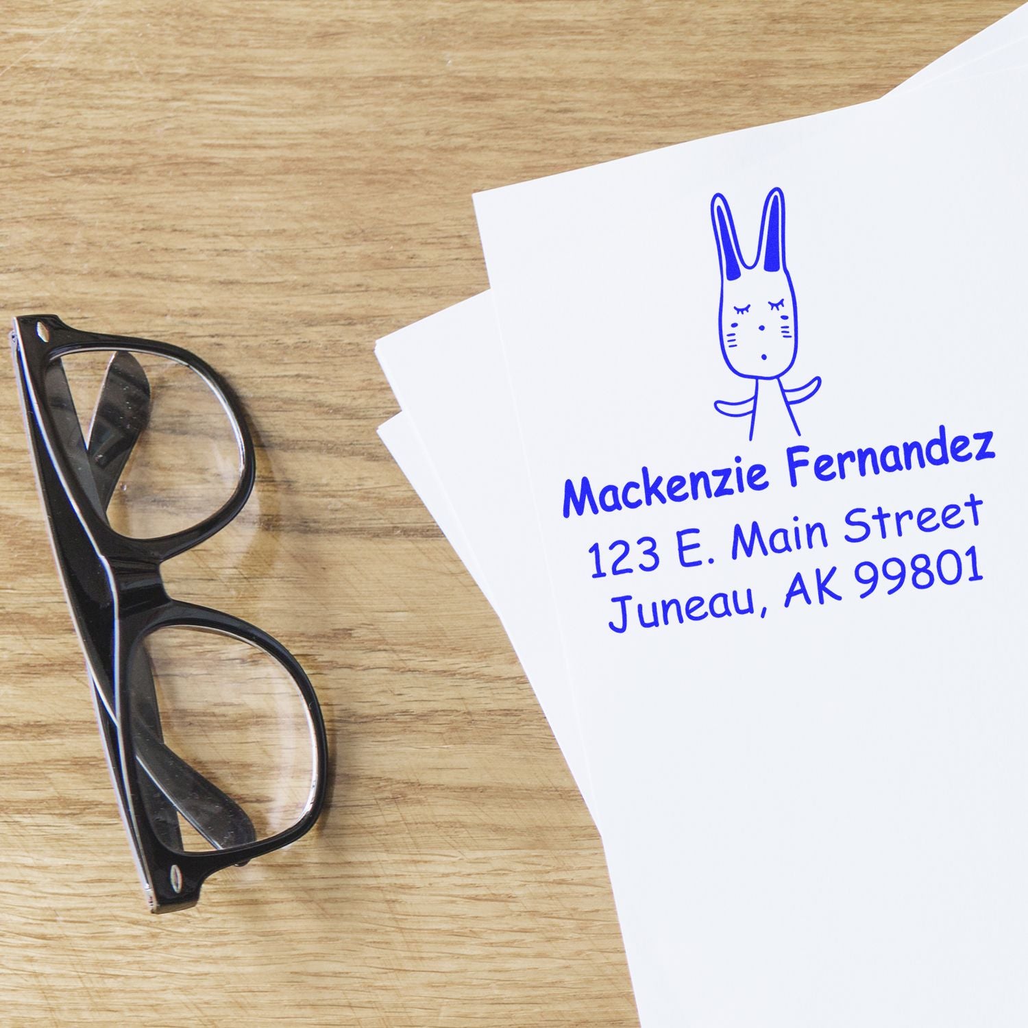 Portable Pre-Inked Hopping Bunny Made To Order Name And Address Stamp on white paper with "Mackenzie Fernandez, 123 E. Main Street, Juneau, AK 99801" in blue ink. Glasses rest nearby. Personalized New Address Stamp design.
