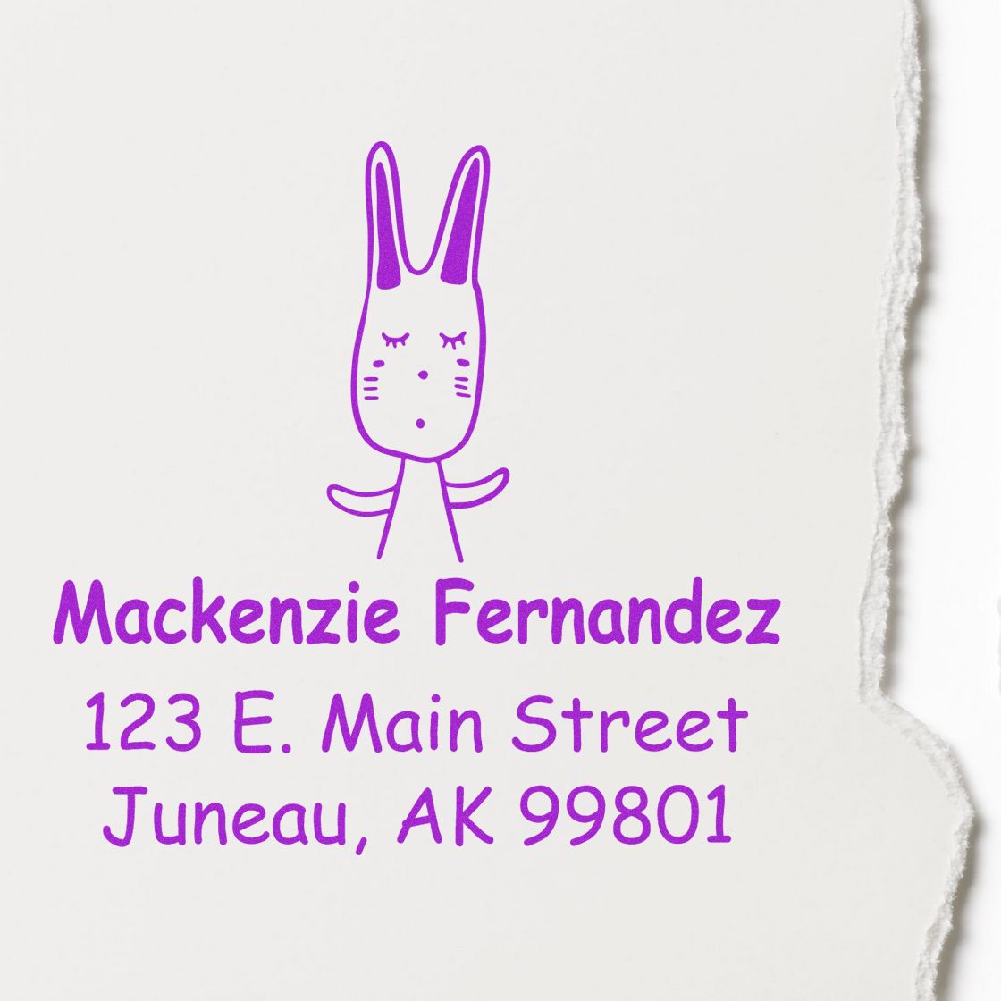 Portable Pre-Inked Hopping Bunny Made To Order Name And Address Stamp featuring a cute bunny design. Personalized Address Stamp displays "Mackenzie Fernandez, 123 E. Main Street, Juneau, AK 99801" in purple ink on white paper with a torn edge.