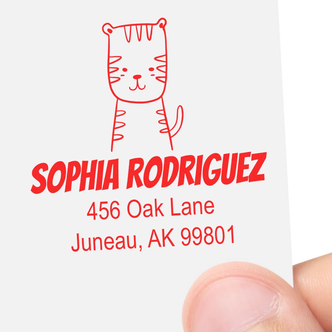 A hand holds a card stamped with a red illustration of a tiger above the name "Sophia Rodriguez" and address "456 Oak Lane, Juneau, AK 99801." The Portable Pre-Inked Terrific Tiger Made To Order Home Address Stamp is a Customized Home Stamper.