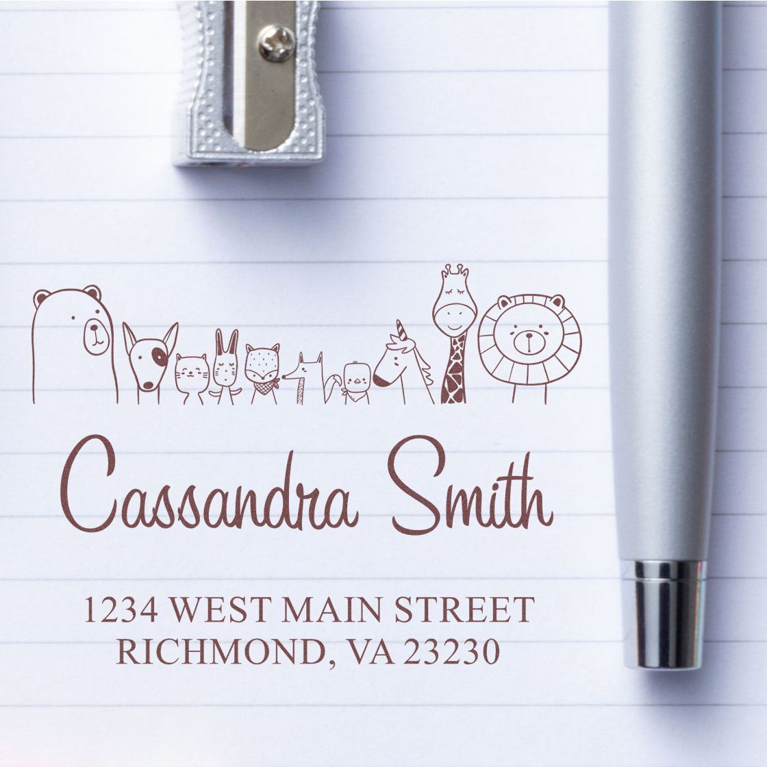 Self-Inking Cartoon Animal Zany Zoo Personalized Return Address Stamper on lined paper. Features custom-made mailing stamp with whimsical animal designs and the name "Cassandra Smith" with an address in Richmond, VA. Silver pen and sharpener nearby.
