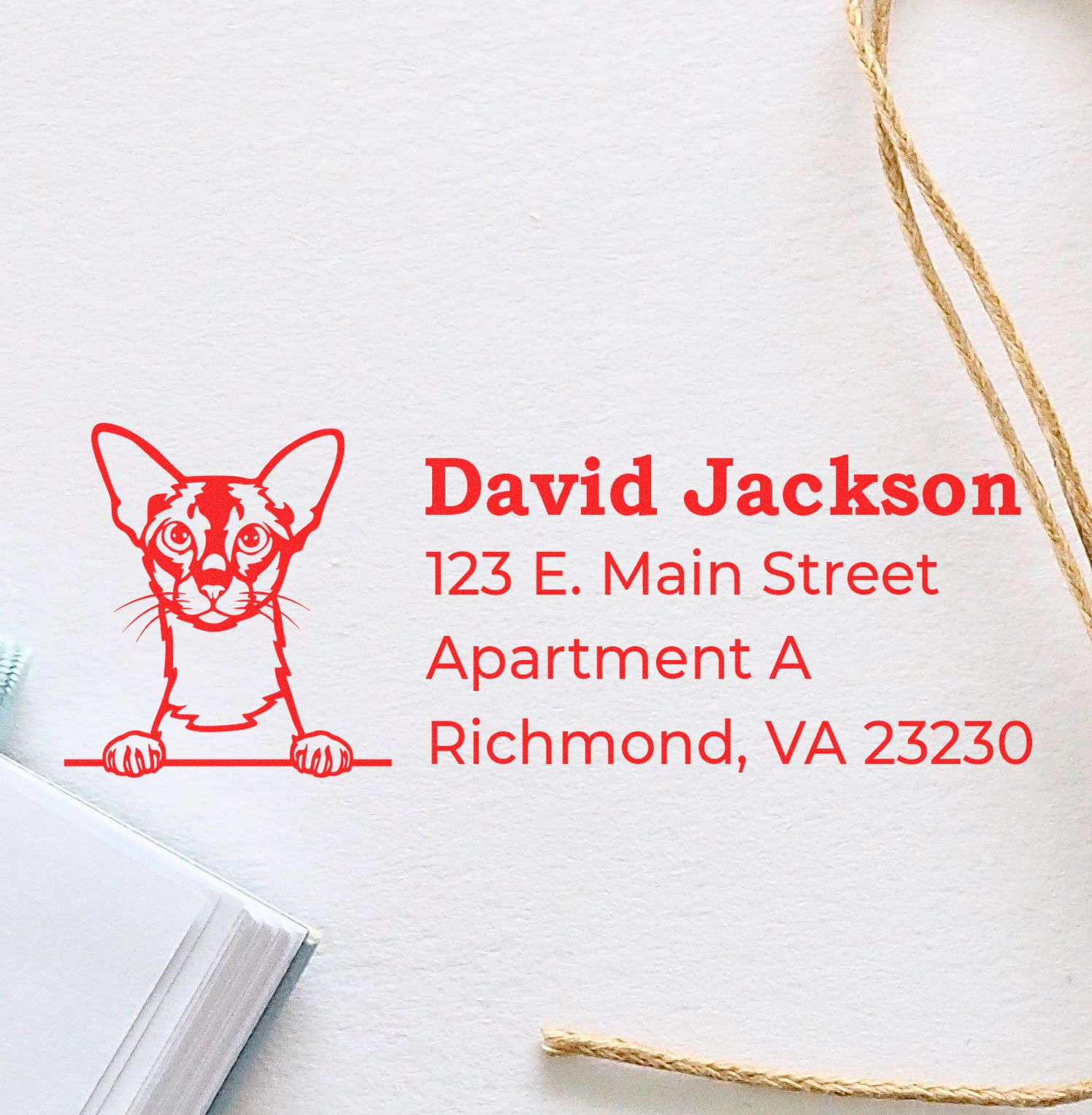 Wood Handle Colorpoint Shorthair Cat Address Stamp on white paper, featuring a red cat illustration and sample address text. Nearby are twine and a notebook, showcasing its elegant design.