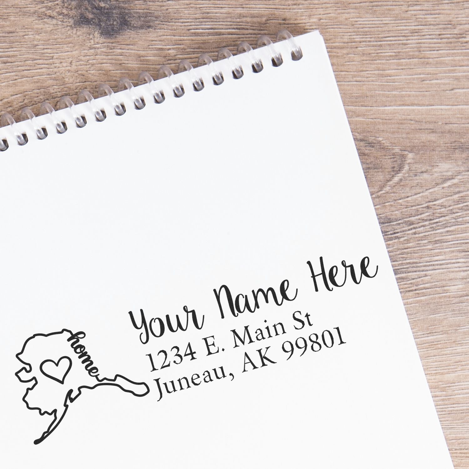 Wood Handle Alaska Custom Address Rubber Stamp on a notepad, featuring a map outline with a heart and the text Your Name Here, 1234 E. Main St, Juneau, AK 99801 in elegant script.
