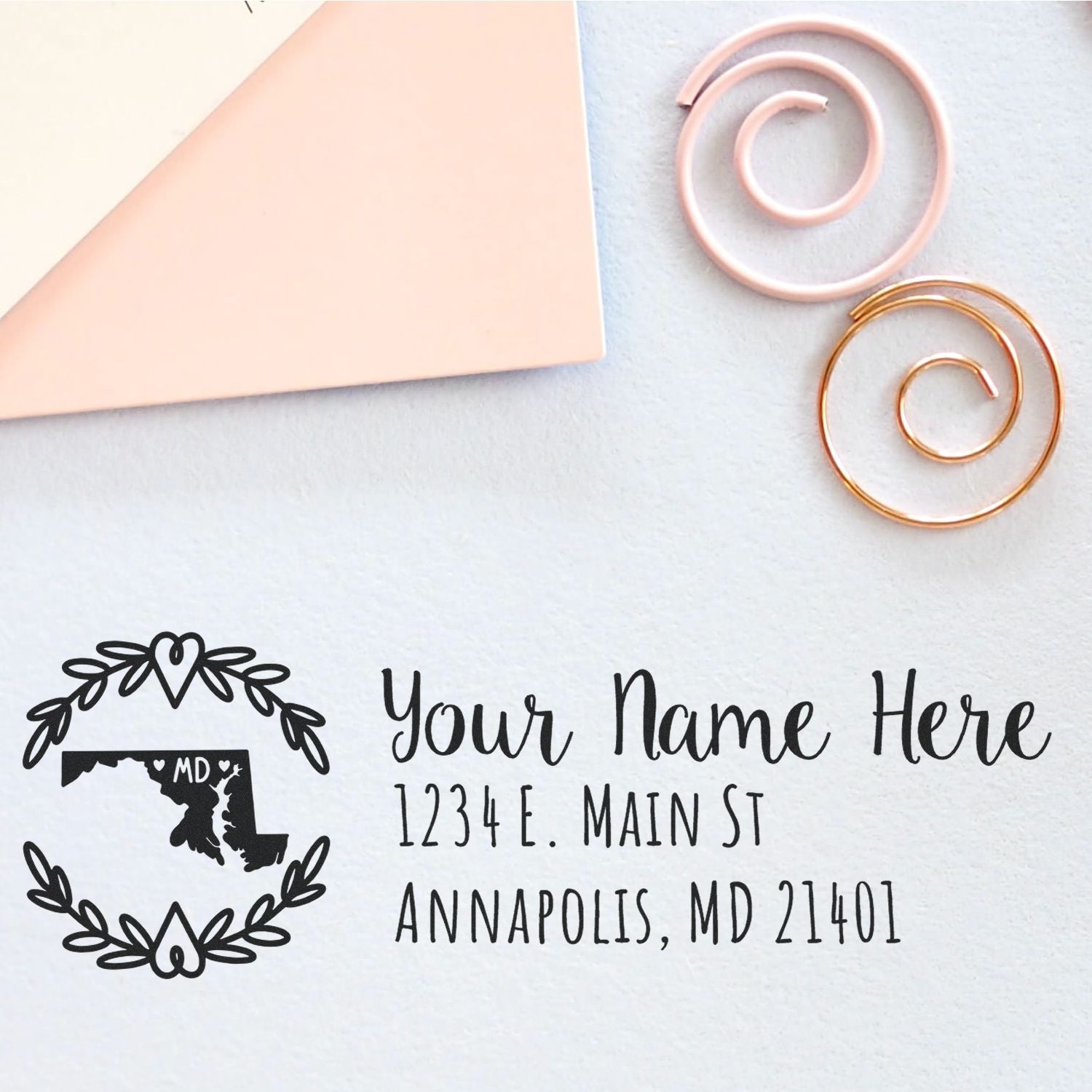 Custom black address stamp with Maryland wreath seal design, pale pink envelope, and rose gold paper clips on white background.