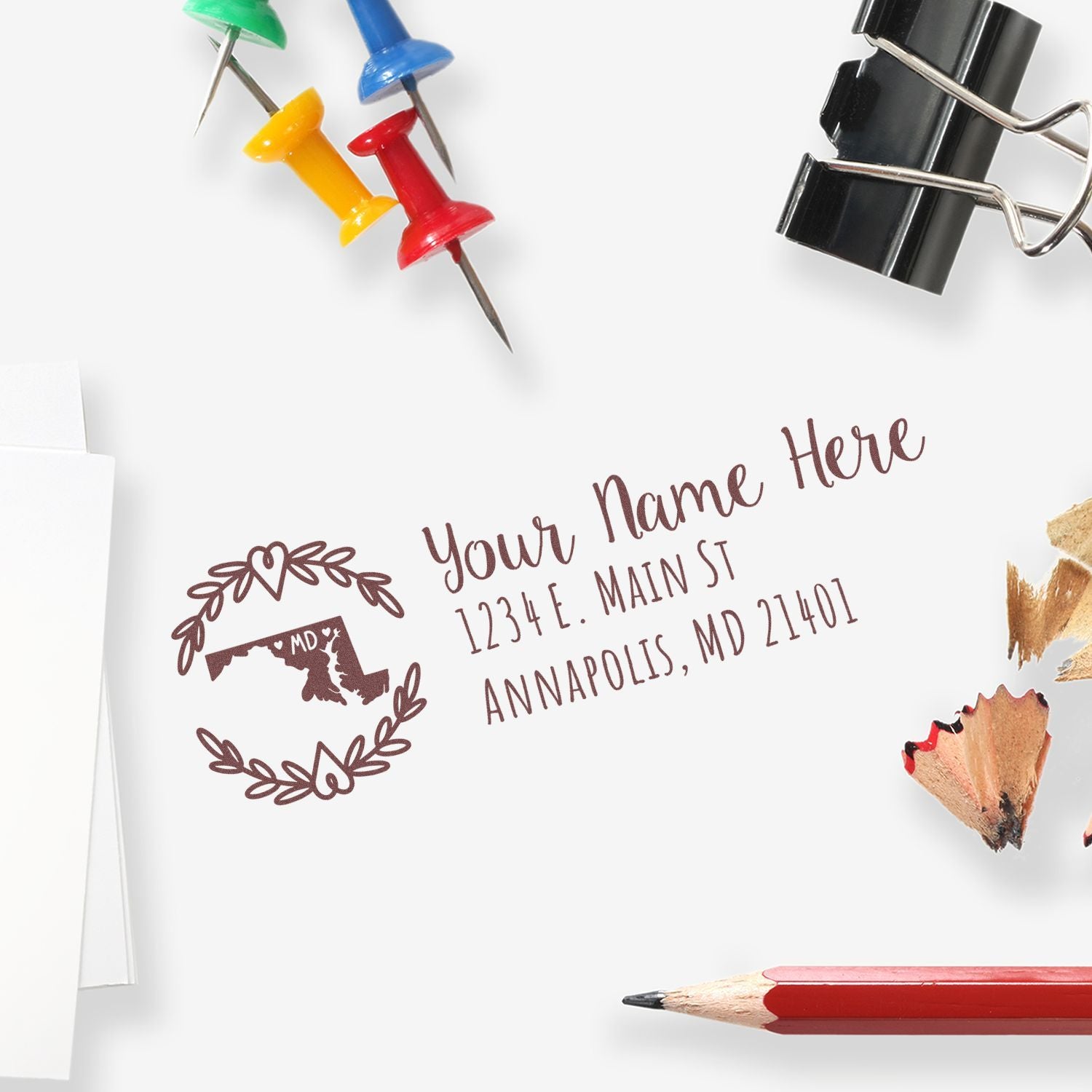 Custom return address stamp and seal with brown Maryland emblem, shown with colorful pushpins, binder clip, and red pencil.