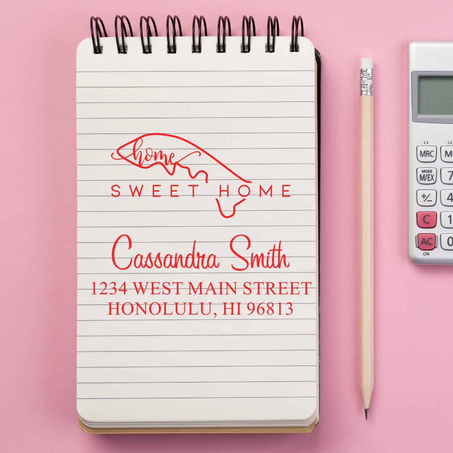 Red custom home address stamp and seal on white spiral notebook, pink background, with pencil and calculator.