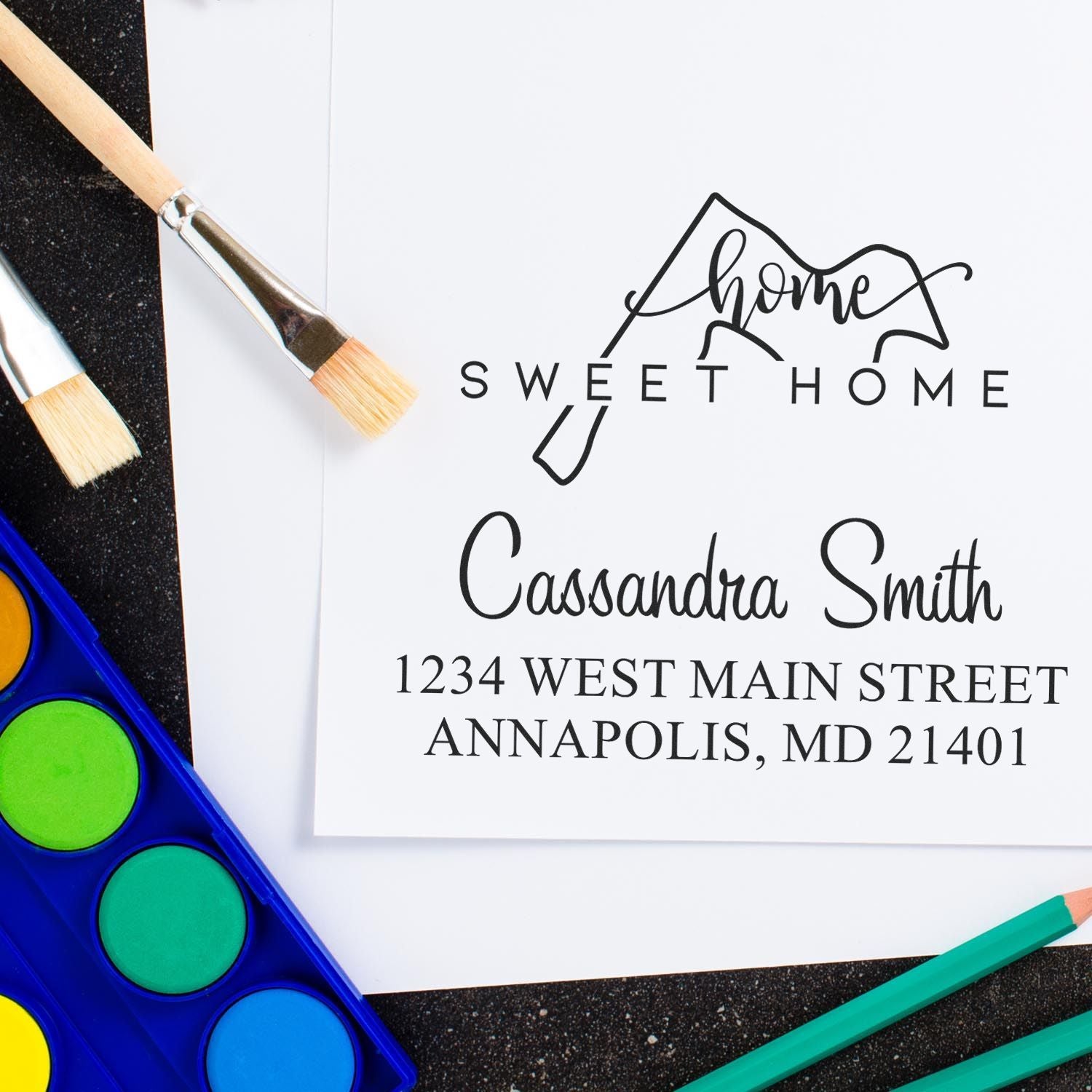 Custom home sweet home address stamp and seal on white paper, with black script text, paintbrushes, and bright watercolor paints.