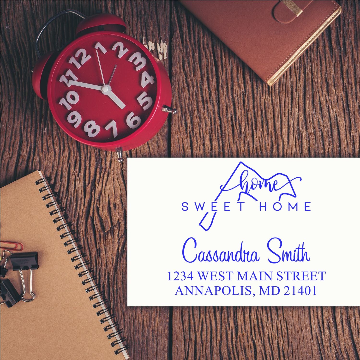 Red alarm clock with brown notebook beside white custom address stamp and seal card, blue Sweet Home text on wood grain.