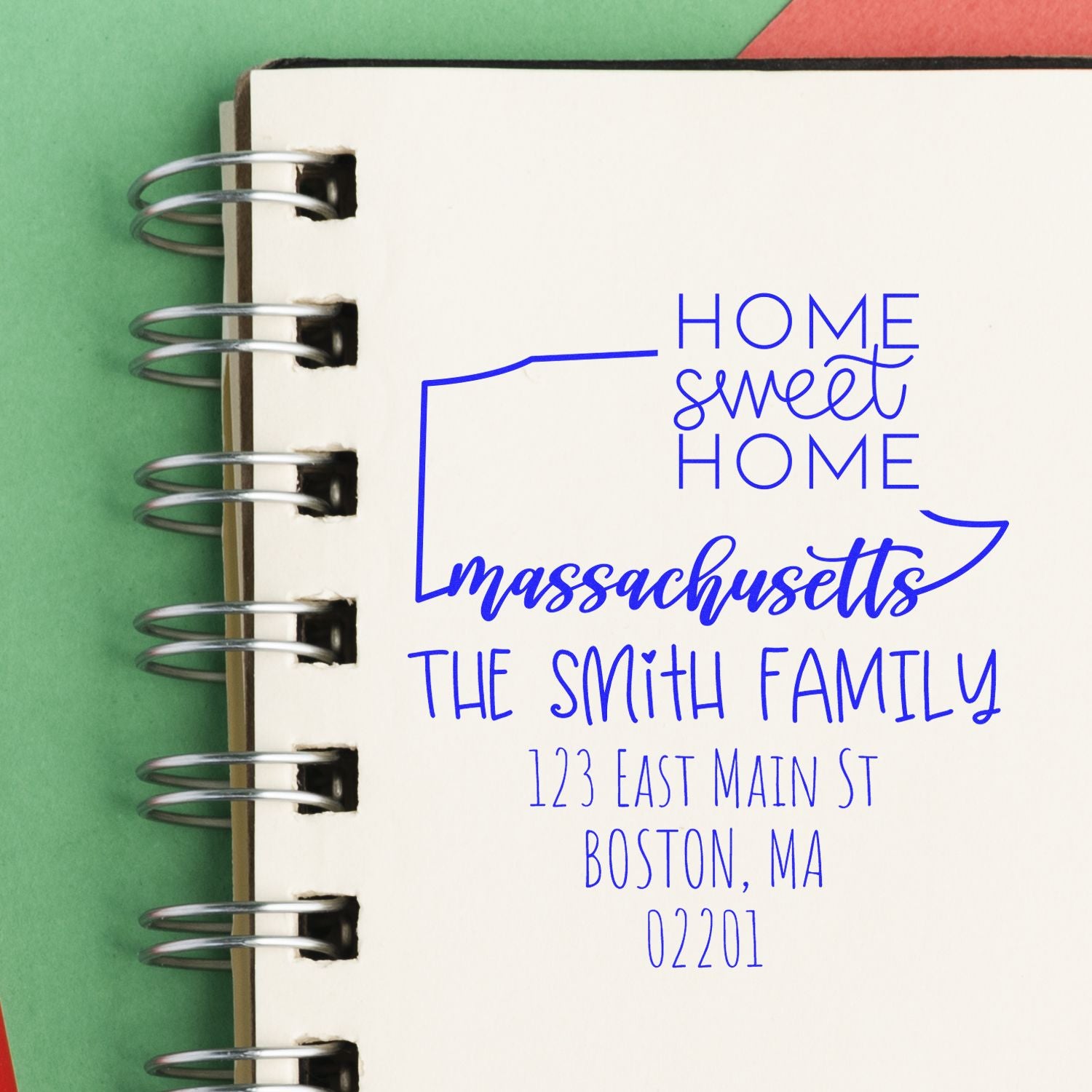 Blue custom address stamp reading Home Sweet Home, Massachusetts on a white spiral notebook with green background.