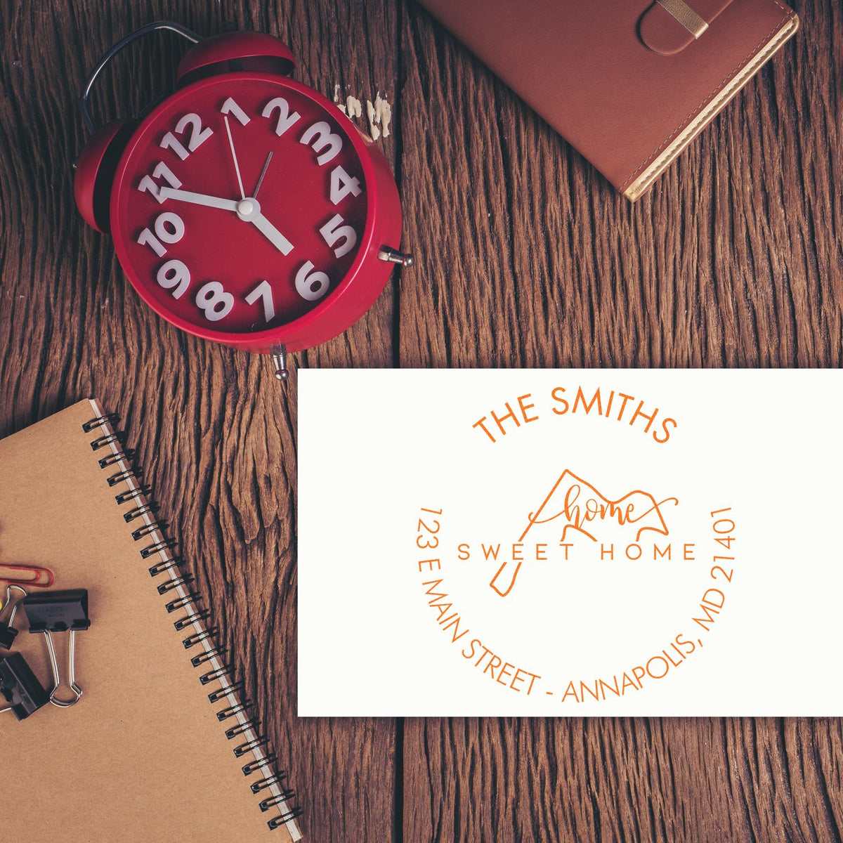 Red alarm clock beside brown notebooks and leather planner with orange custom home address stamp seal on wood desk.