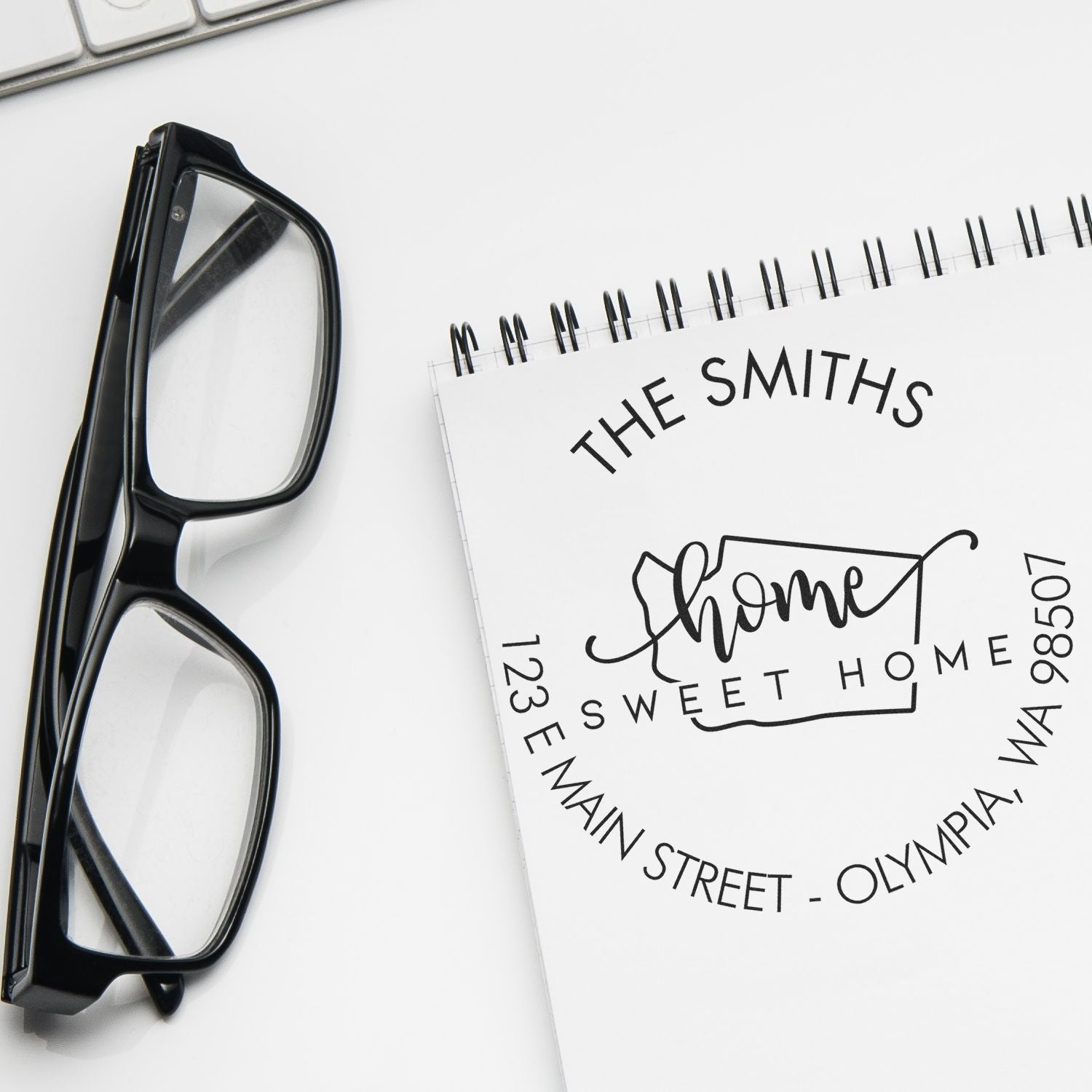 Black eyeglasses beside a white spiral notebook with a custom address stamp seal reading The Smiths Home Sweet Home.