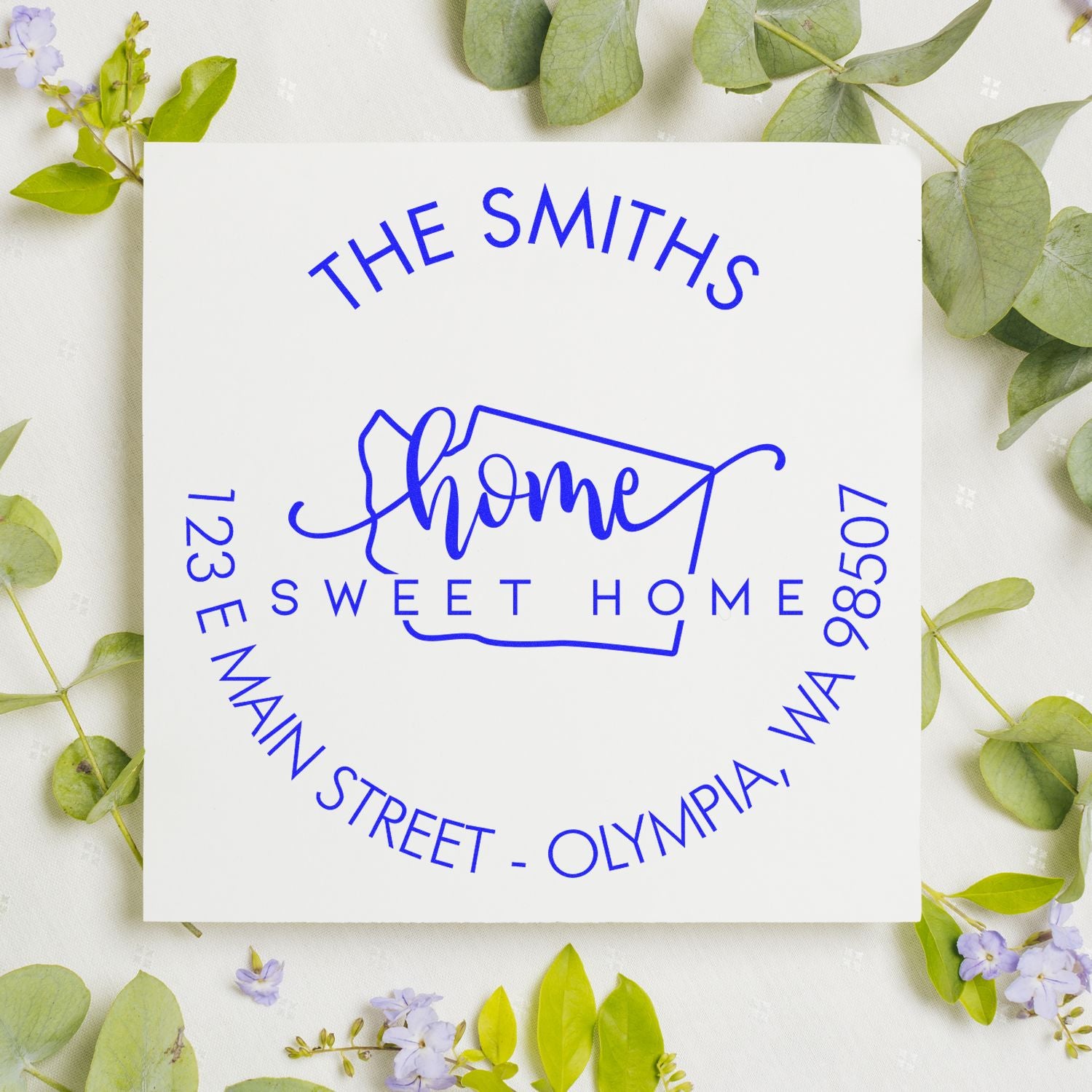 Blue custom home address stamp seal on white card, surrounded by green leaves and small purple flowers