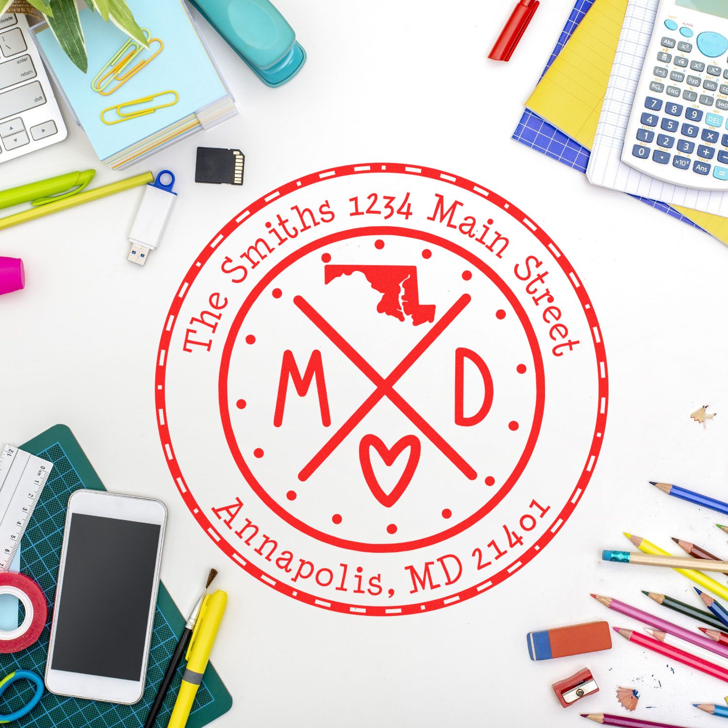 Red custom stamp seal with Maryland outline, M-D monogram and heart, on a white desk with colorful office supplies.