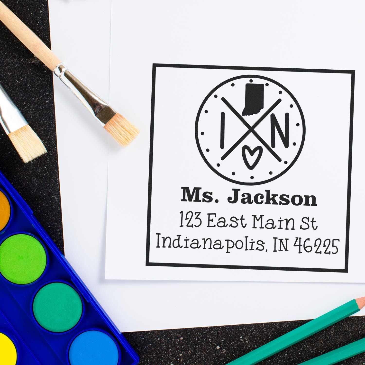 Custom black teacher stamp and seal with Ms. Jackson address, surrounded by paintbrushes, watercolor palette, and pencils.