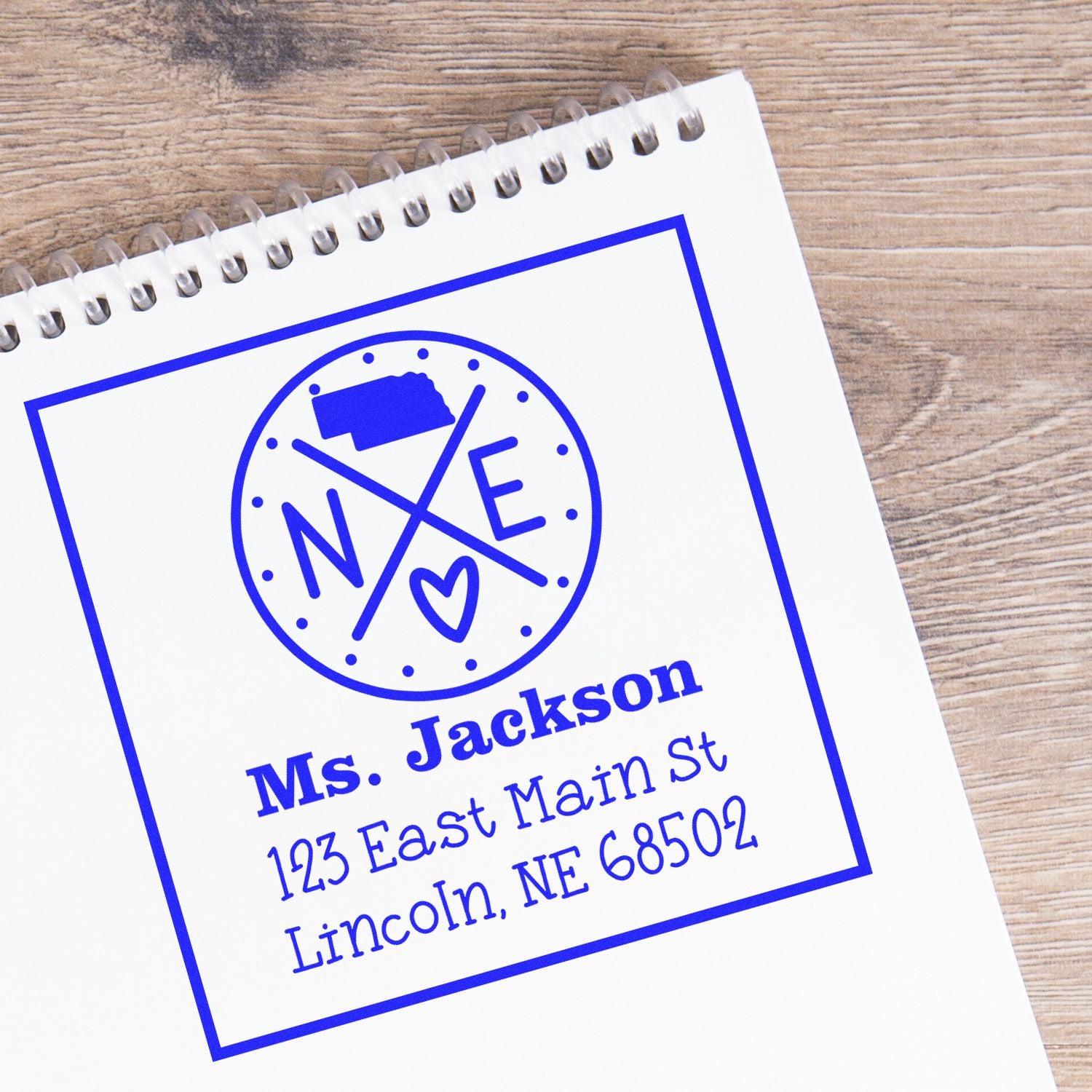 Blue custom self-inking address stamp and seal for Ms. Jackson on a white spiral notebook, with heart logo, on wood table.