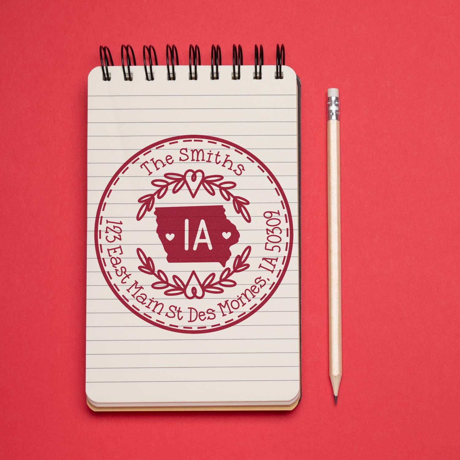 Custom maroon address stamp and seal on lined notebook, with Iowa map, beside a pencil on a red background.
