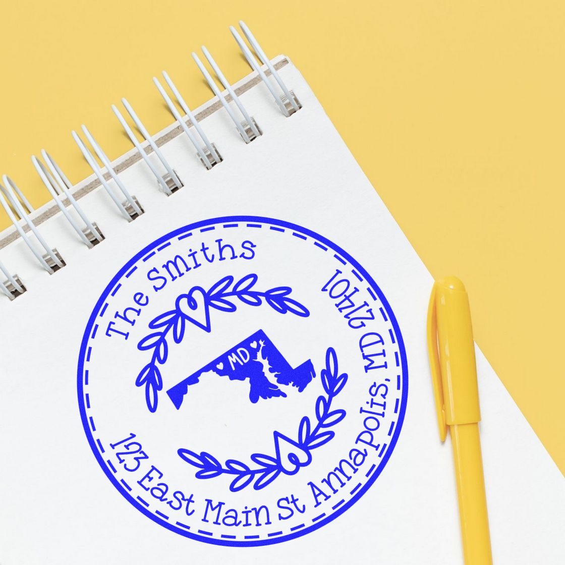 Blue custom stamp seal with wreath and Maryland design on white notebook, beside a yellow pen on yellow background.
