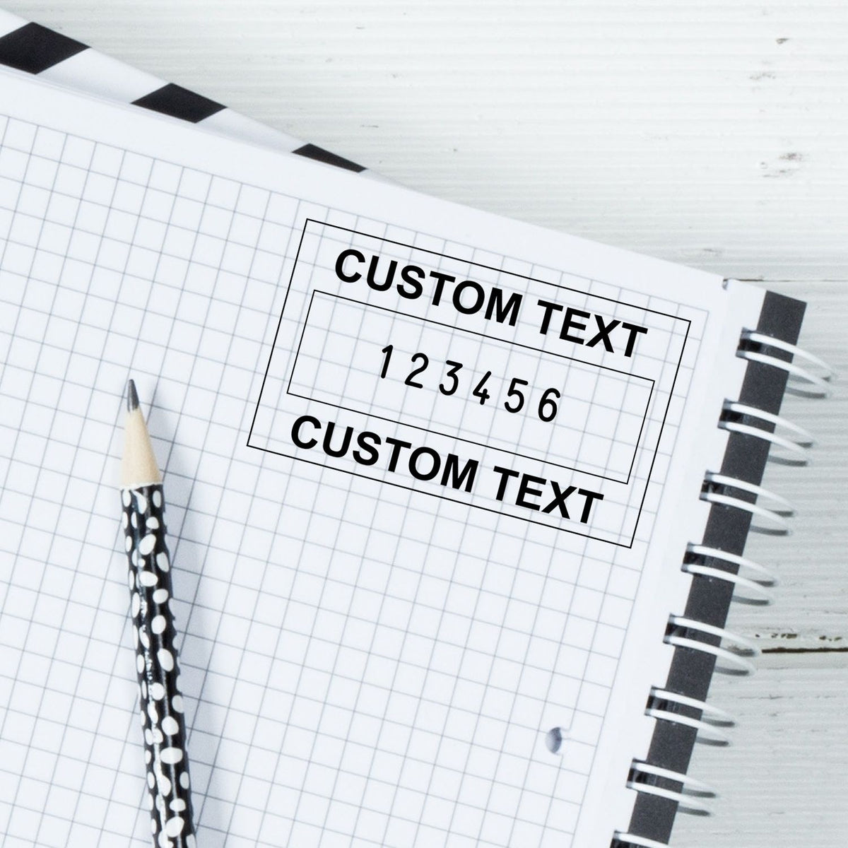 Black custom text stamp seal on white graph notebook beside a black-and-white polka dot pencil.