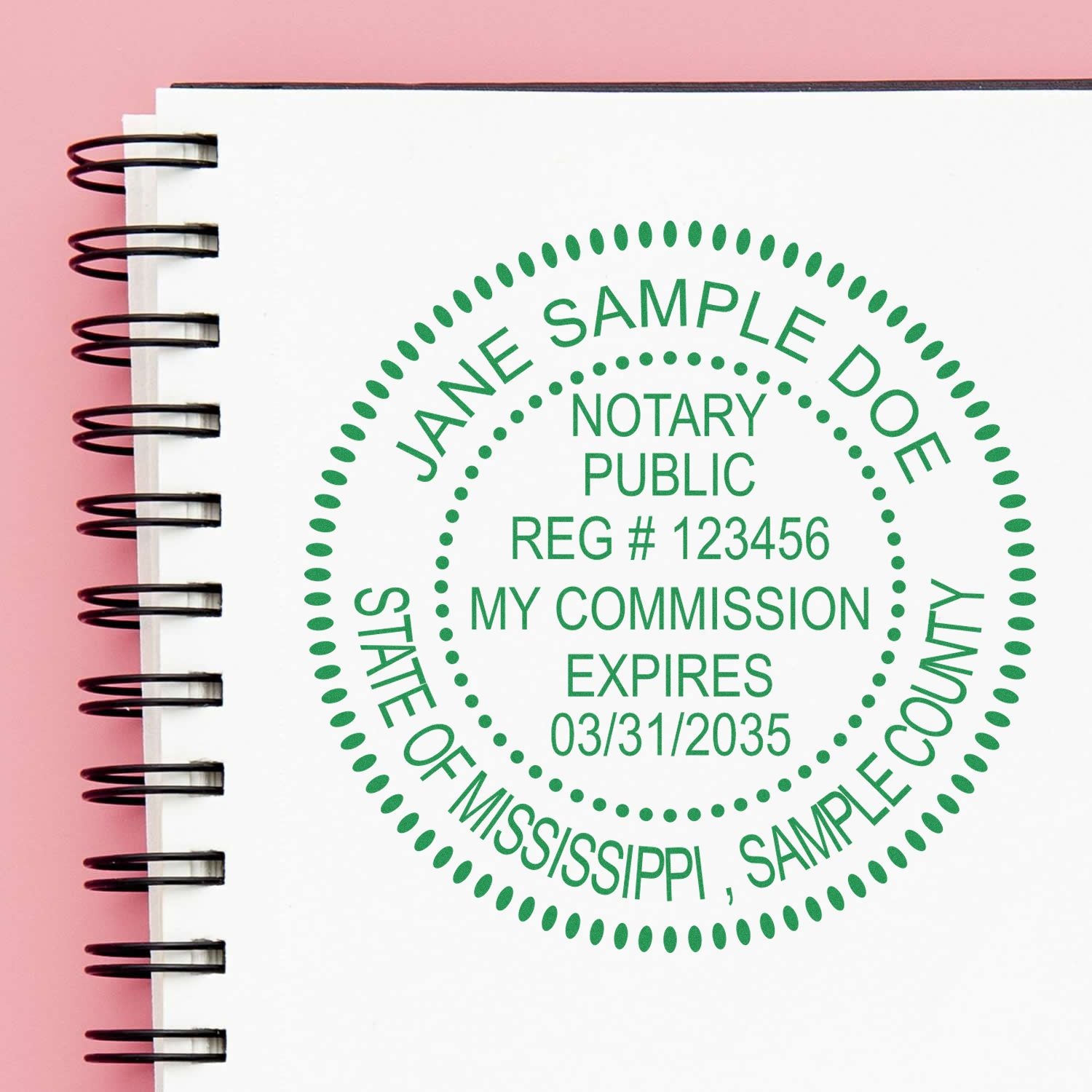 Green notary public stamp seal on a white spiral notebook, with pink background and sample commission details for Mississippi.