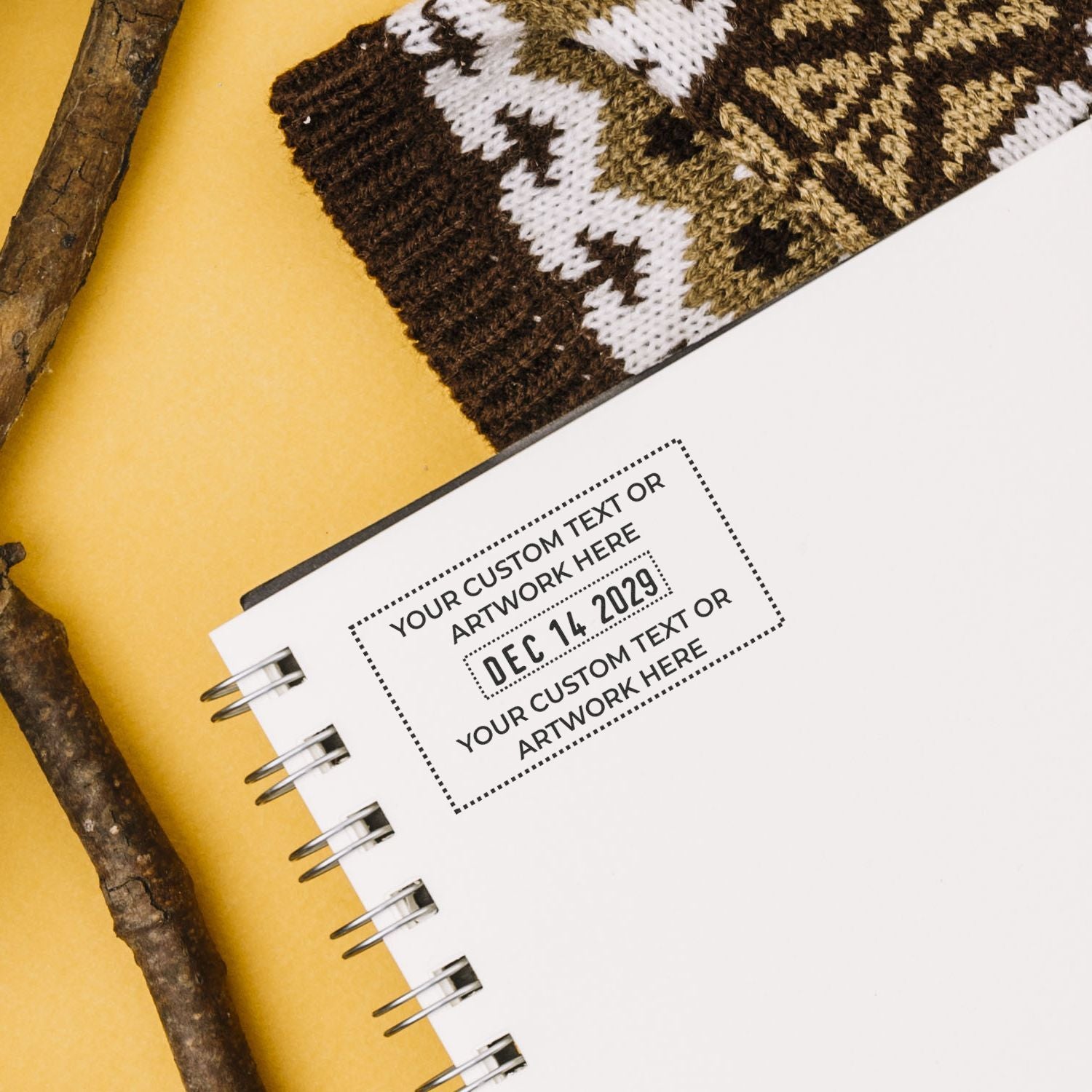 Custom stamp and seal on a white spiral notebook, with brown knit fabric, rustic twigs, and a yellow background.