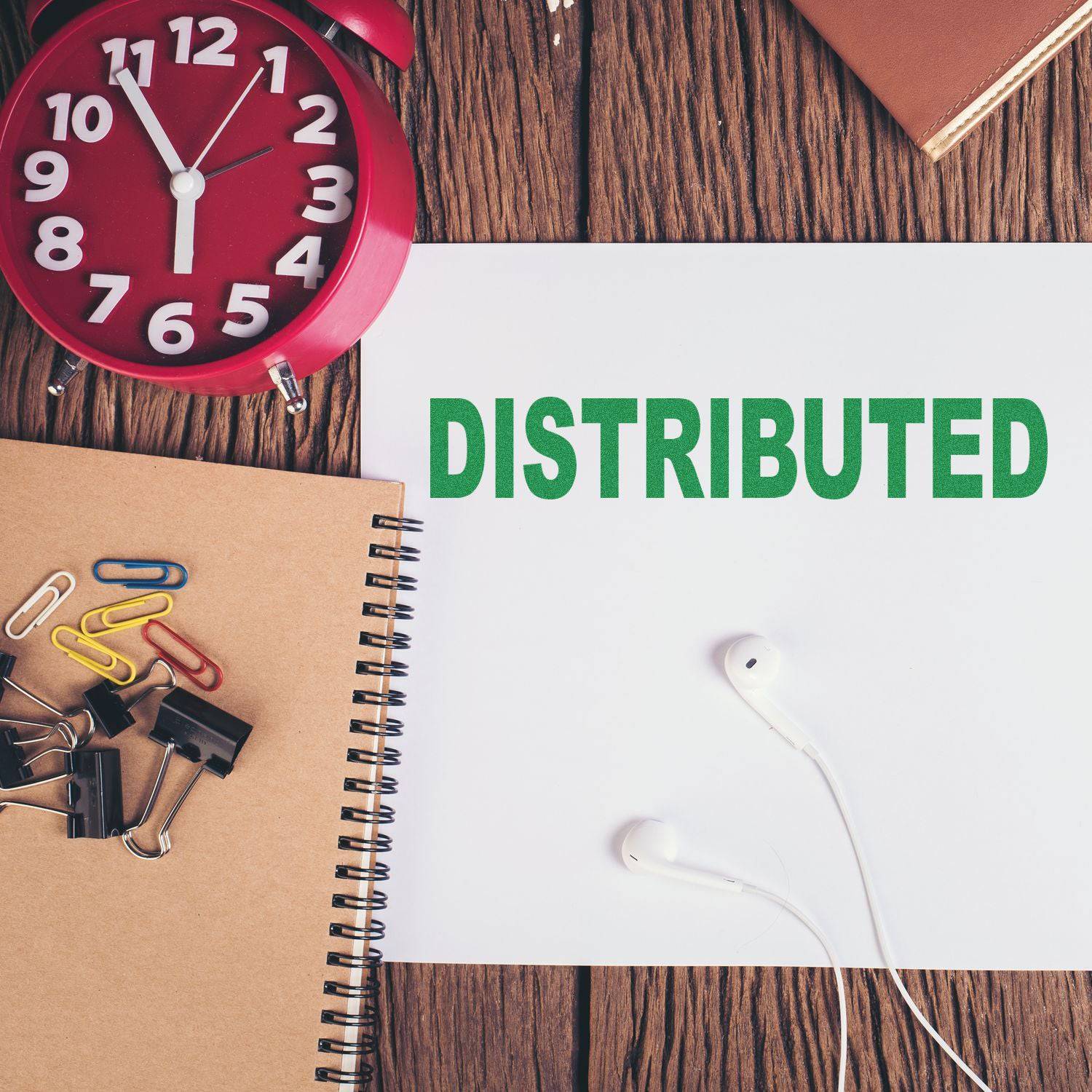 Large Pre-Inked Distributed Stamp on white paper, surrounded by a red clock, notebook, paper clips, binder clips, and earphones.