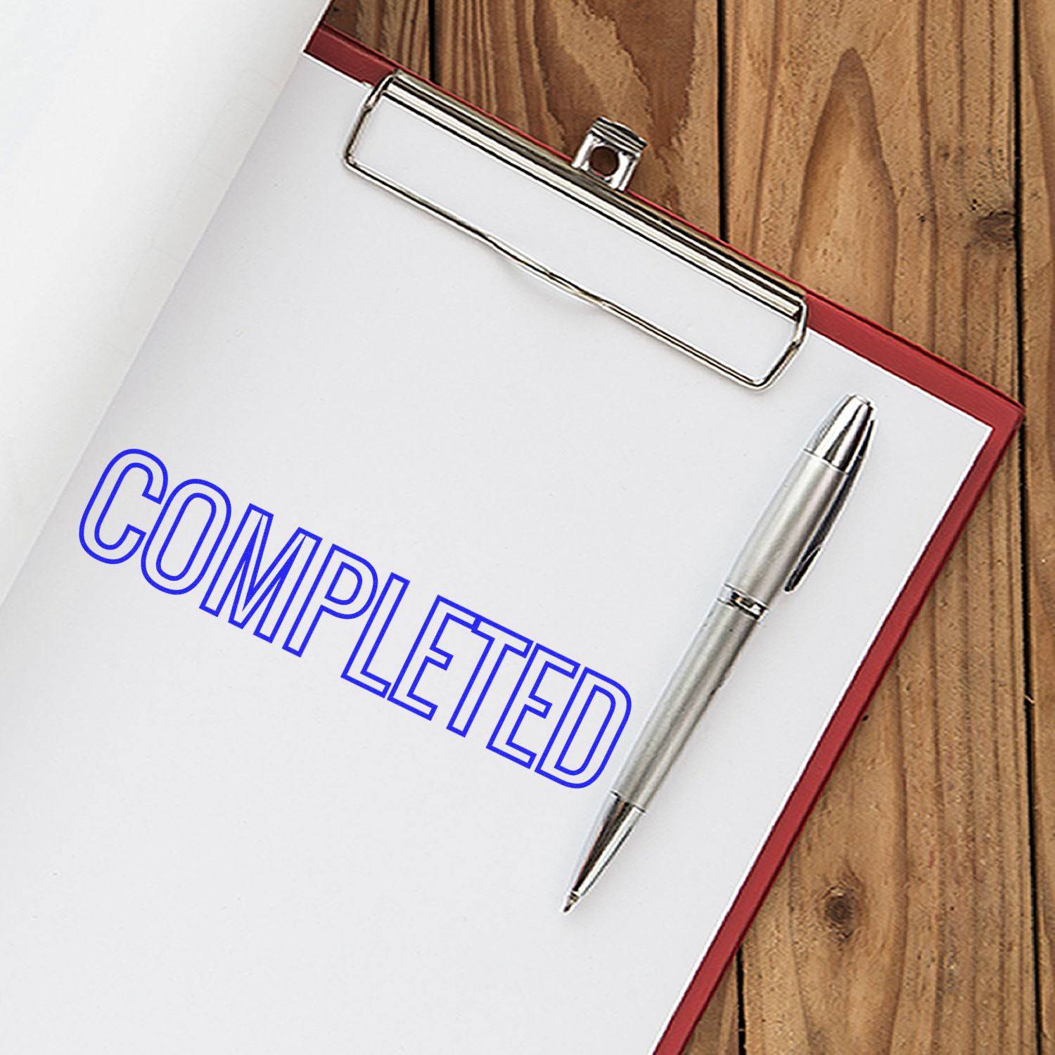 Large Pre-Inked Outline Completed Stamp in blue ink on a white paper clipboard with a silver pen, placed on a wooden surface.