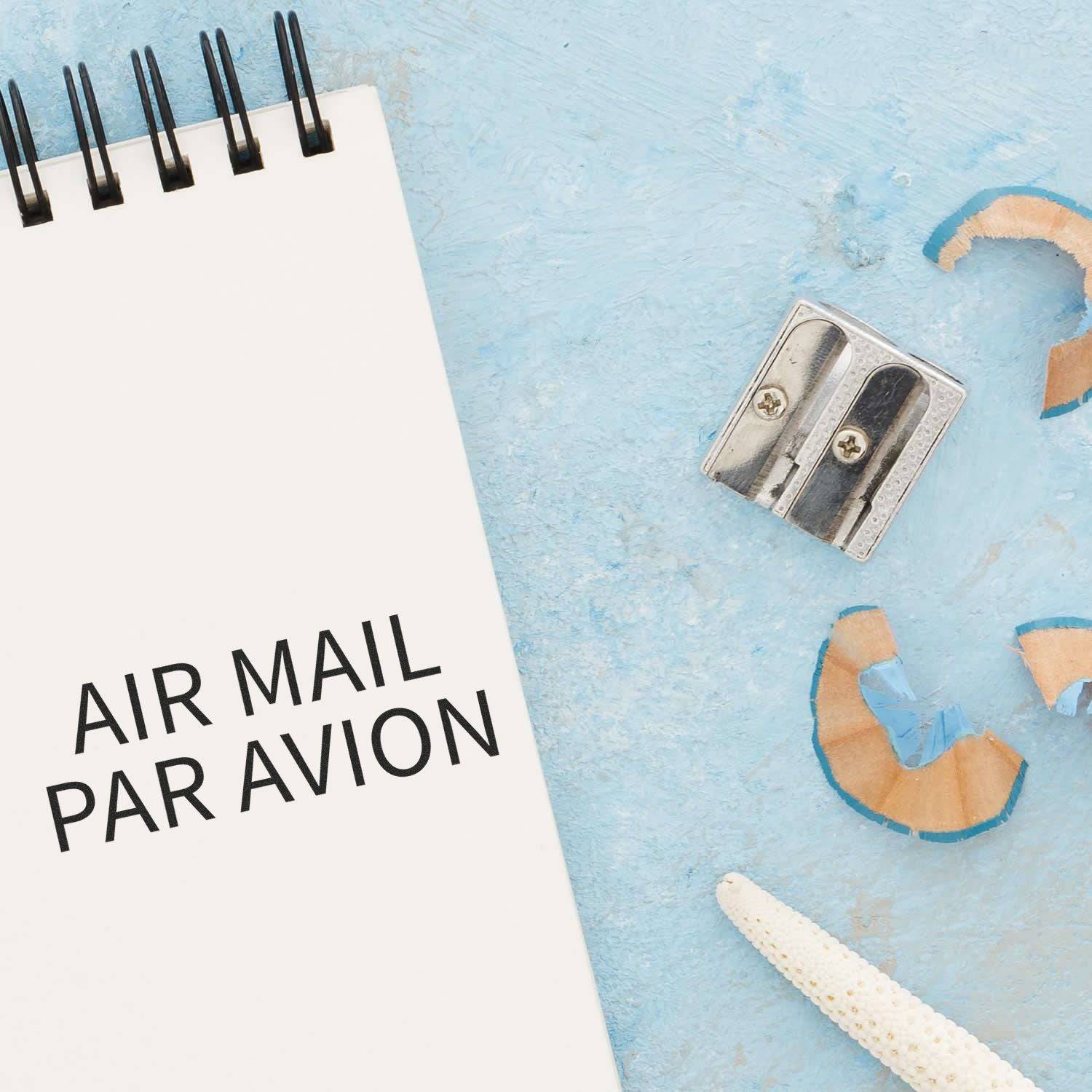 Large Pre-Inked Air Mail Par Avion Stamp on a notepad, with a pencil sharpener and shavings on a light blue background.