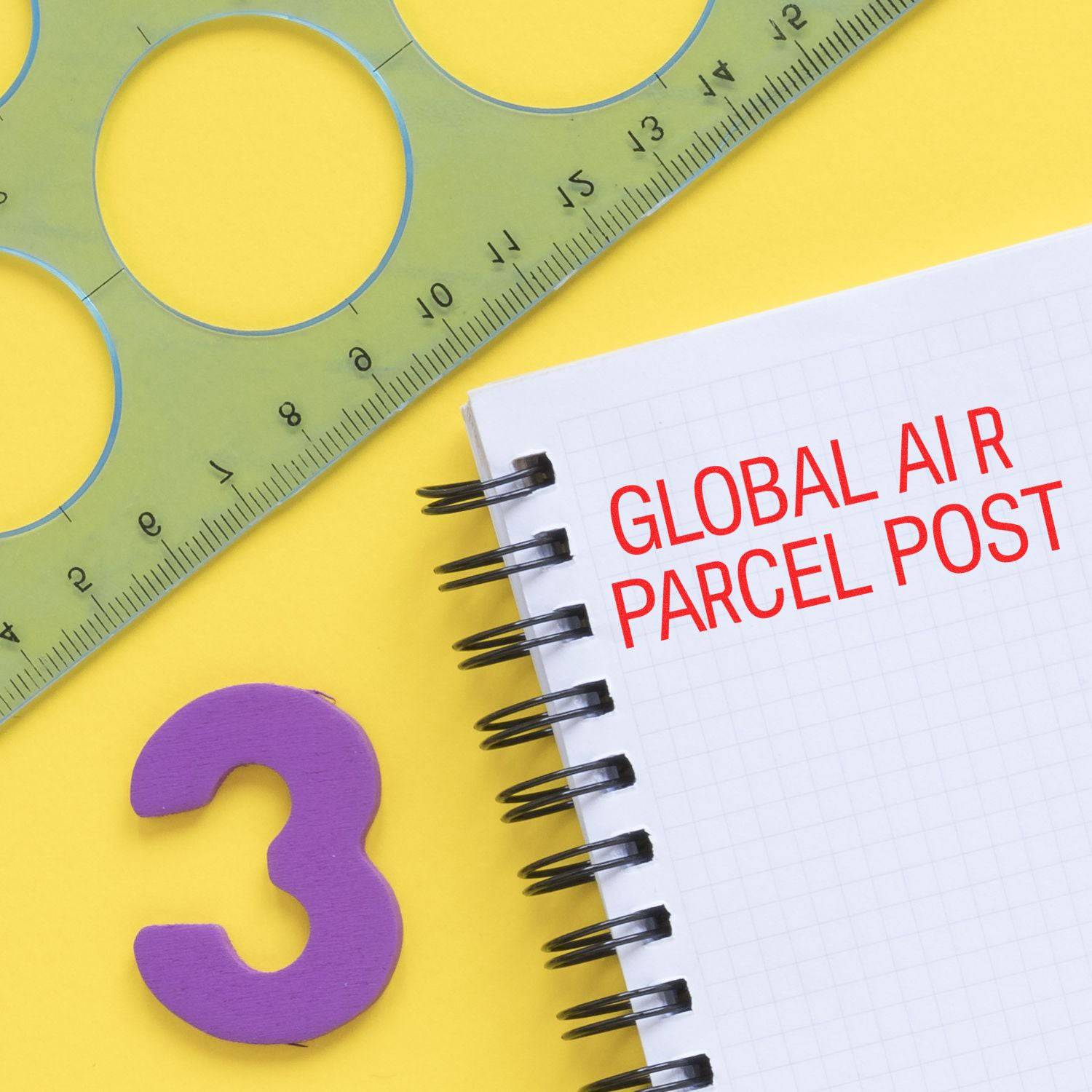 A Large Pre-Inked Global Air Parcel Post Stamp on a spiral notebook with a green ruler and a purple number 3 on a yellow background.