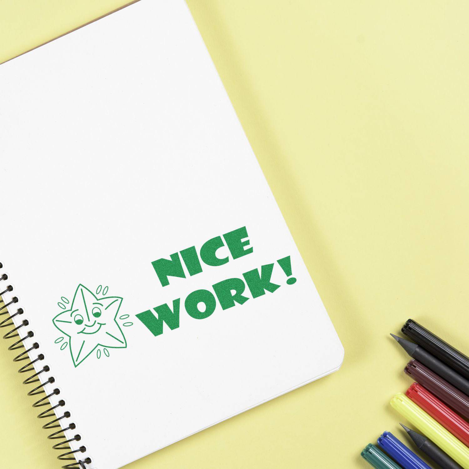 Large Pre-Inked Nice Work Stamp on a notebook with colorful pens on a yellow background.