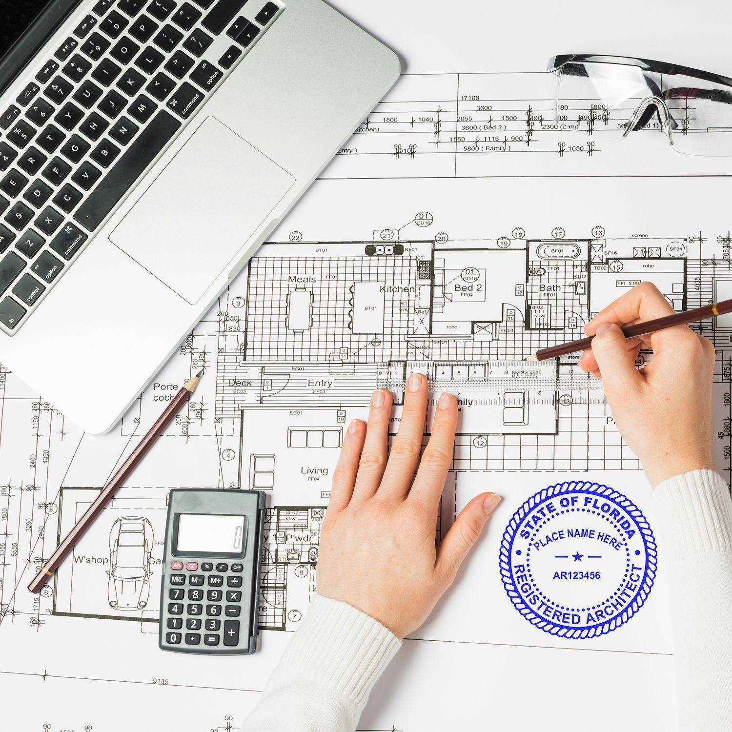 A hand using the Self-Inking Architect Stamp of Seal on a detailed architectural blueprint, with a laptop, calculator, and glasses nearby on the desk.