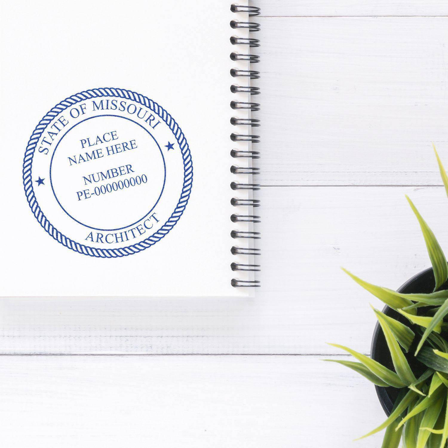 Xstamper Architect Pre-Inked Rubber Stamp of Seal used on a white spiral notebook, displaying a professional seal with 'State of Missouri Architect' text. A green plant is placed beside the notebook.