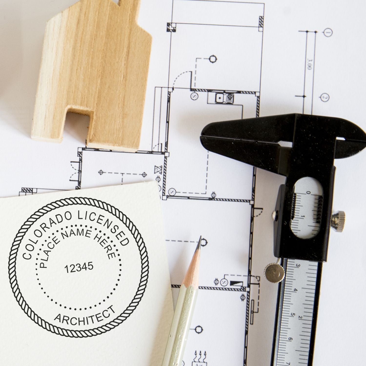 Colorado Architect Seal Stamp in use on architectural plans, with a pencil, caliper, and wooden house model nearby.