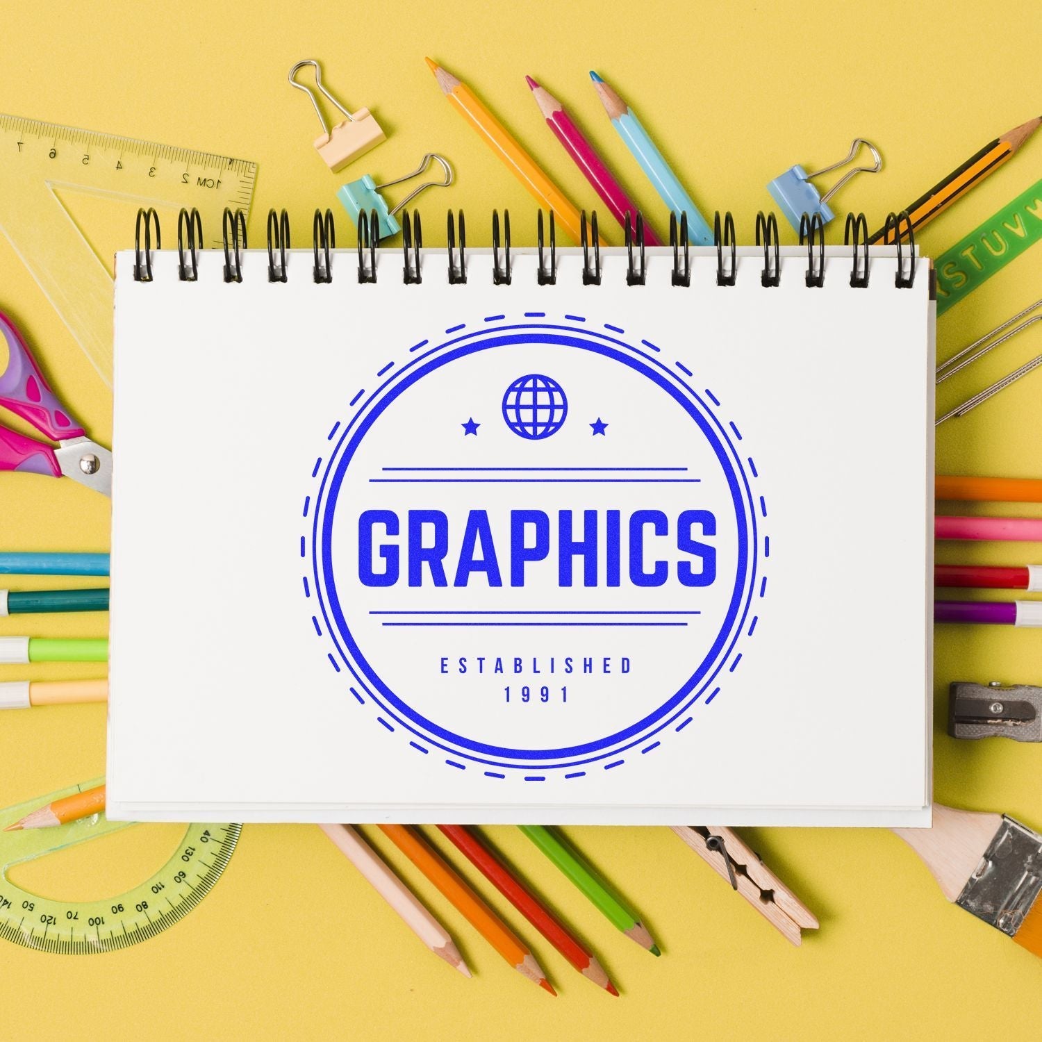 A Custom Self Inking Stamp Trodat 46050 Size 2 Diameter is used to stamp a GRAPHICS logo on a sketchbook surrounded by colorful stationery.
