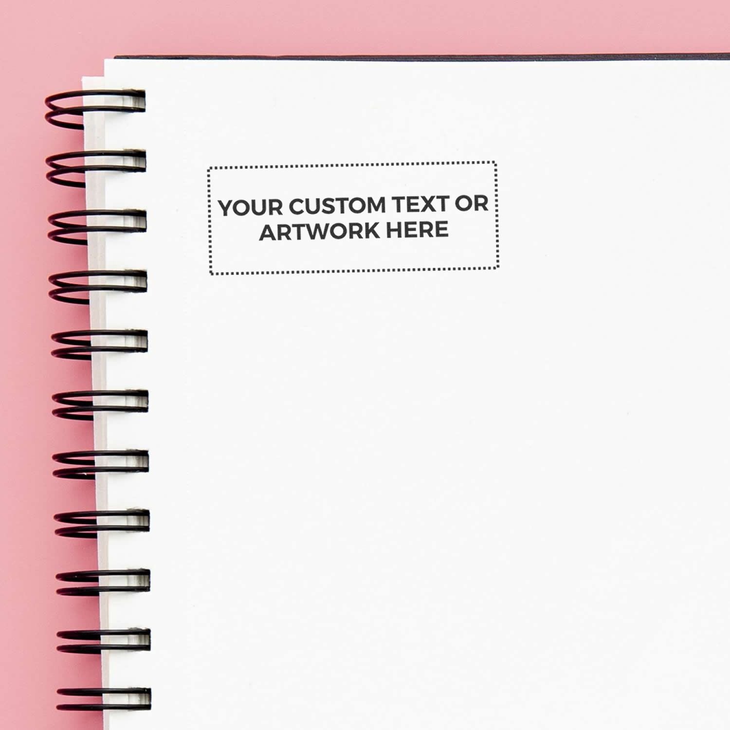 Custom Self Inking Stamp Trodat 4913 Size 7/8 x 2-3/8 imprint on a white spiral notebook with a pink background.