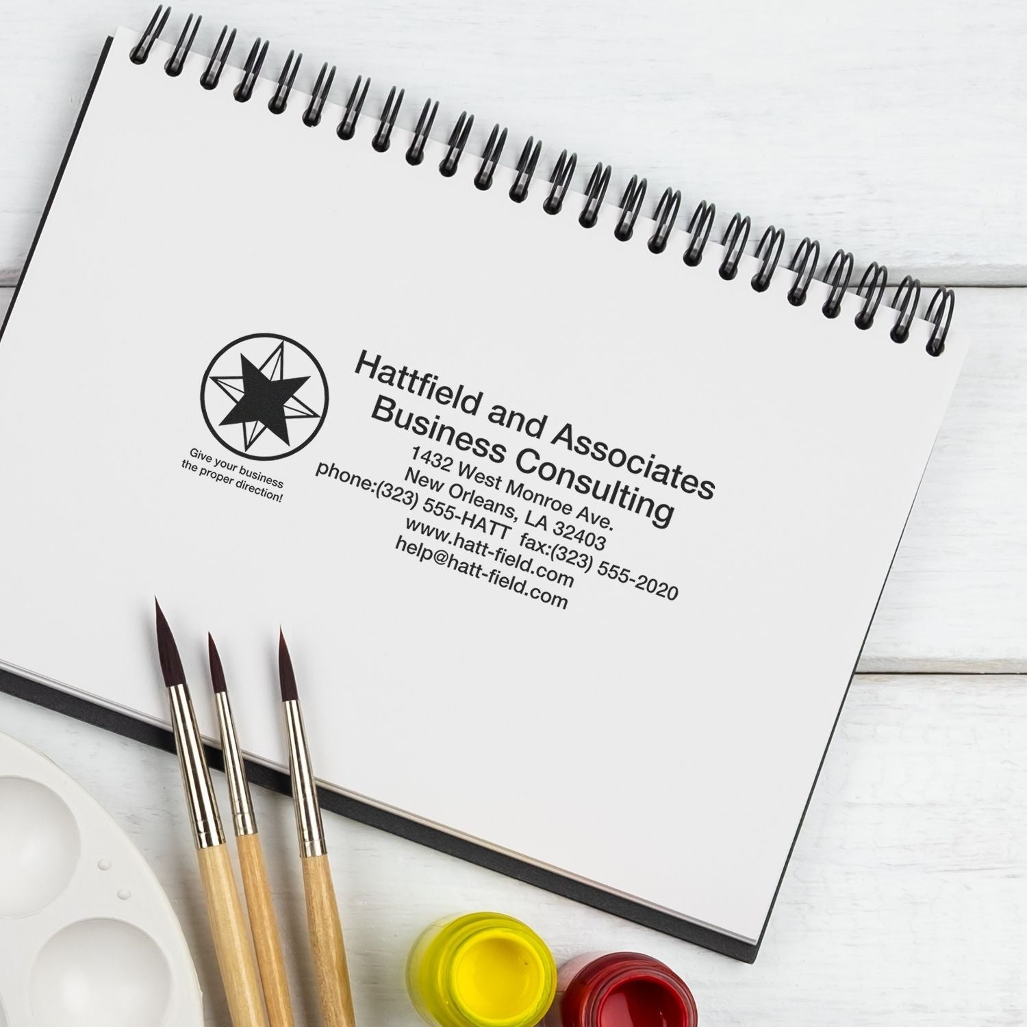 Custom Self Inking Stamp Trodat 5212 Size 2-3/4 x 4-1/2 used on a spiral notebook with business consulting details, next to paintbrushes.
