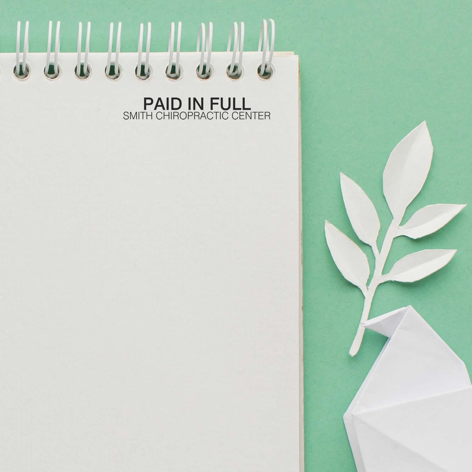 A Custom Rubber Stamp Size 1 x 3 is used on a spiral notebook with PAID IN FULL and SMITH CHIROPRACTIC CENTER stamped on a green background.
