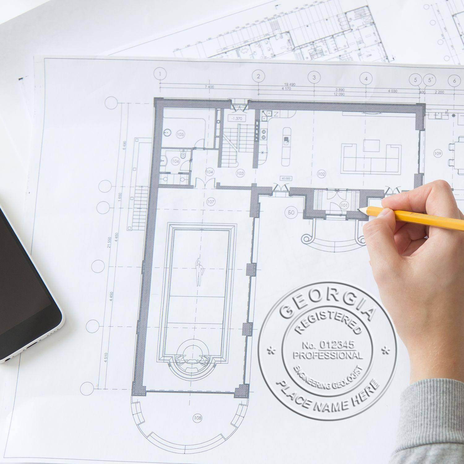 A hand holding a pencil over architectural blueprints with an embossed seal impression reading Engineering Geologist Chrome Gift Seal Embosser on the paper. A smartphone is placed on the left side.