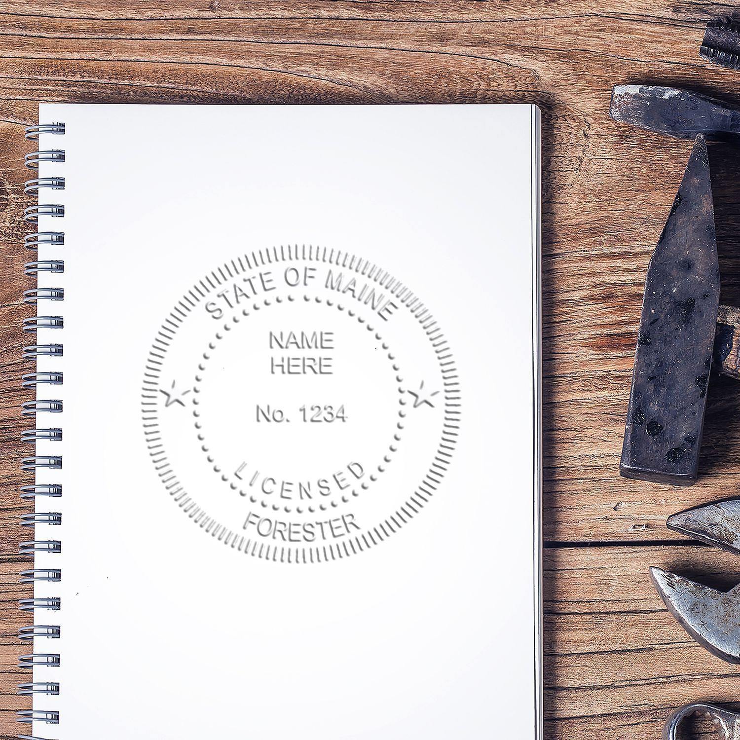 Forester Desk Seal Embosser imprint on a white notebook, placed on a wooden desk with tools nearby.