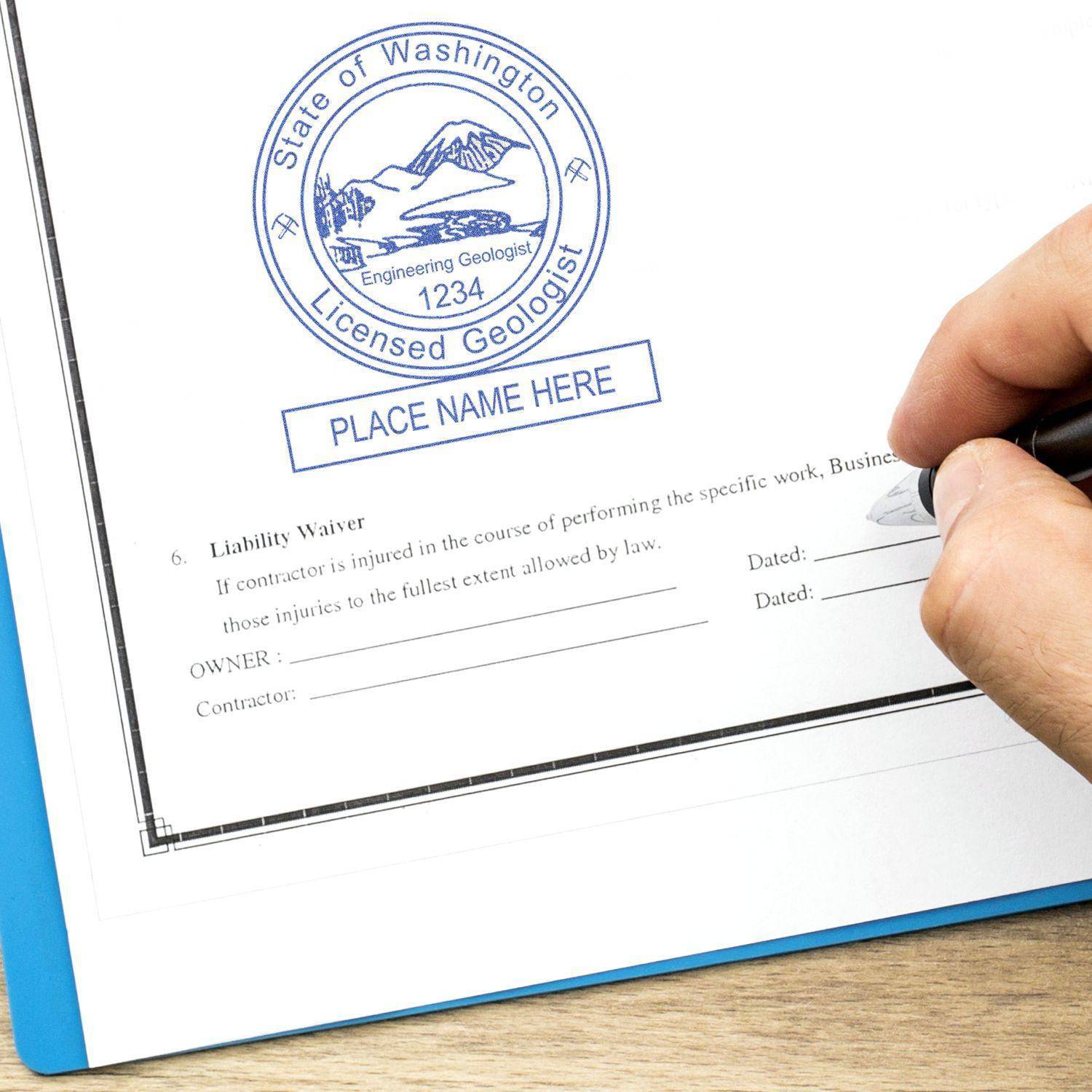 A hand using the Engineering Geologist MaxLight Pre-Inked Rubber Stamp of Seal on a document, displaying a blue circular seal with State of Washington Licensed Geologist and space for a name and date.