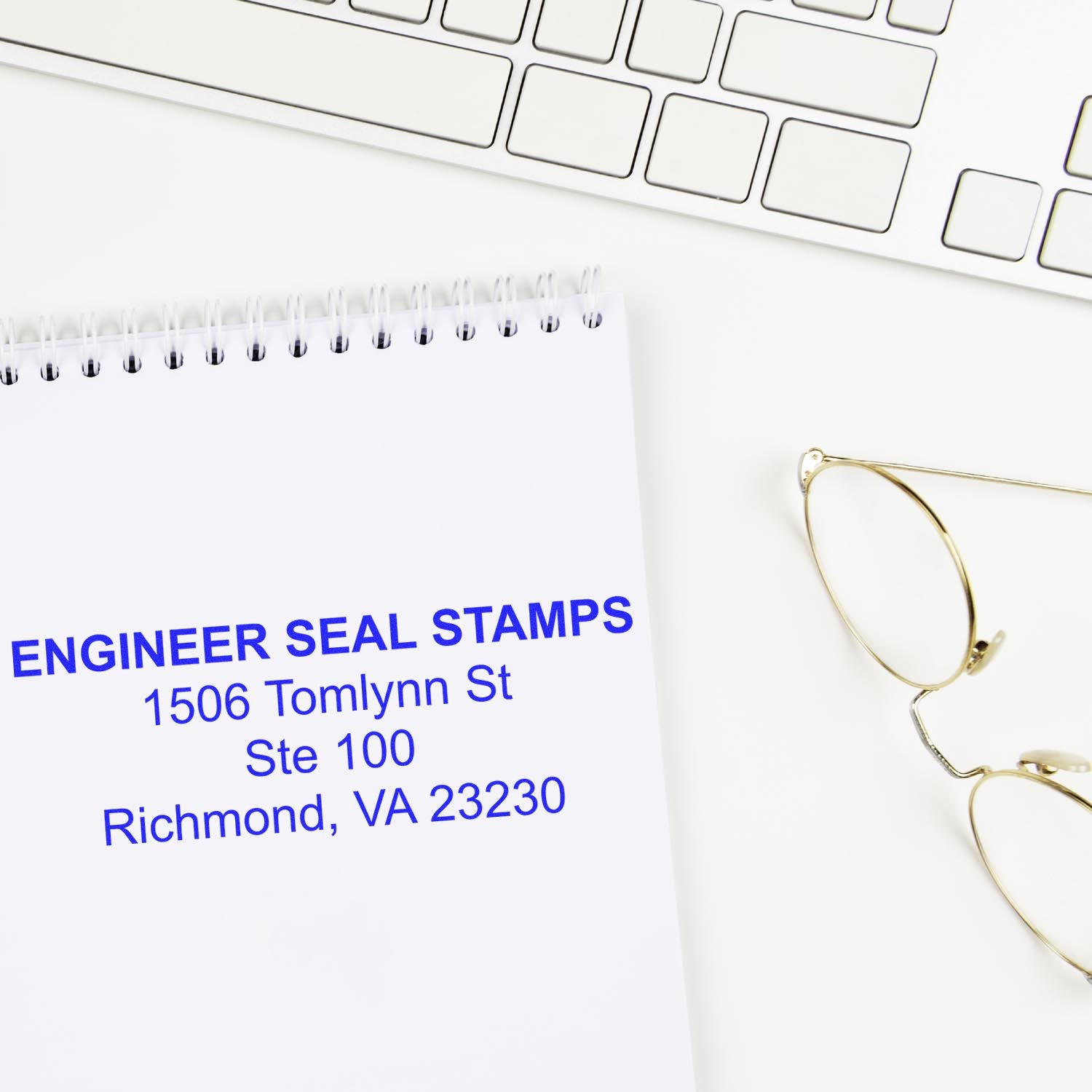 A 4 Line Self Inking Address Stamp imprint on a notepad, placed next to a keyboard and glasses. The address reads ENGINEER SEAL STAMPS, 1506 Tomlynn St, Ste 100, Richmond, VA 23230 .