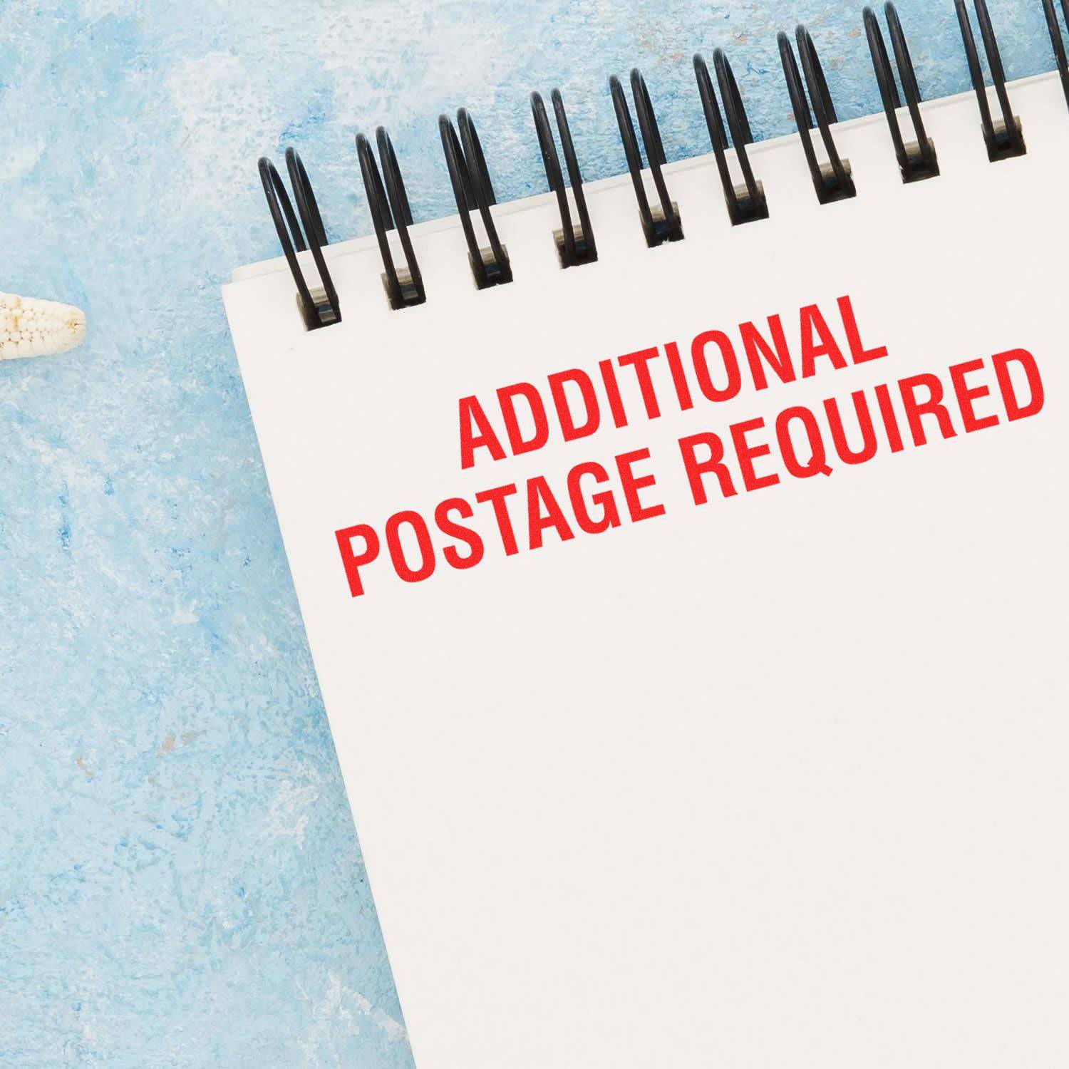 Large Self Inking Additional Postage Required Stamp in red ink on a white notepad with a blue background.