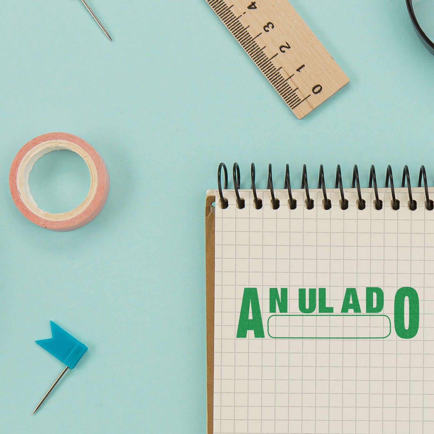 Large Self Inking Anulado Stamp in green ink on a spiral notebook, surrounded by a ruler, tape, and a blue flag pin on a light blue background.