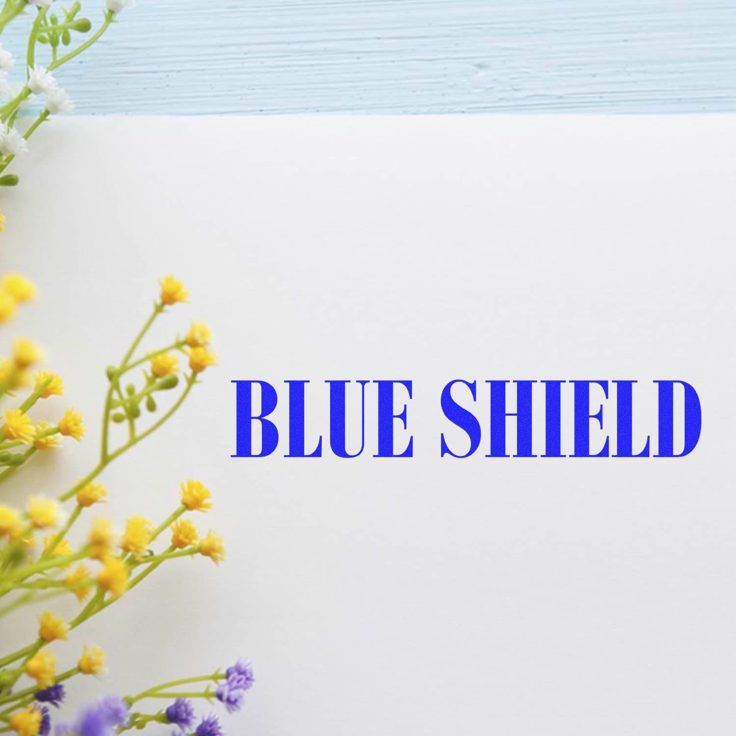 Large Self Inking Blue Shield Stamp imprint on white paper with yellow and purple flowers on the side.