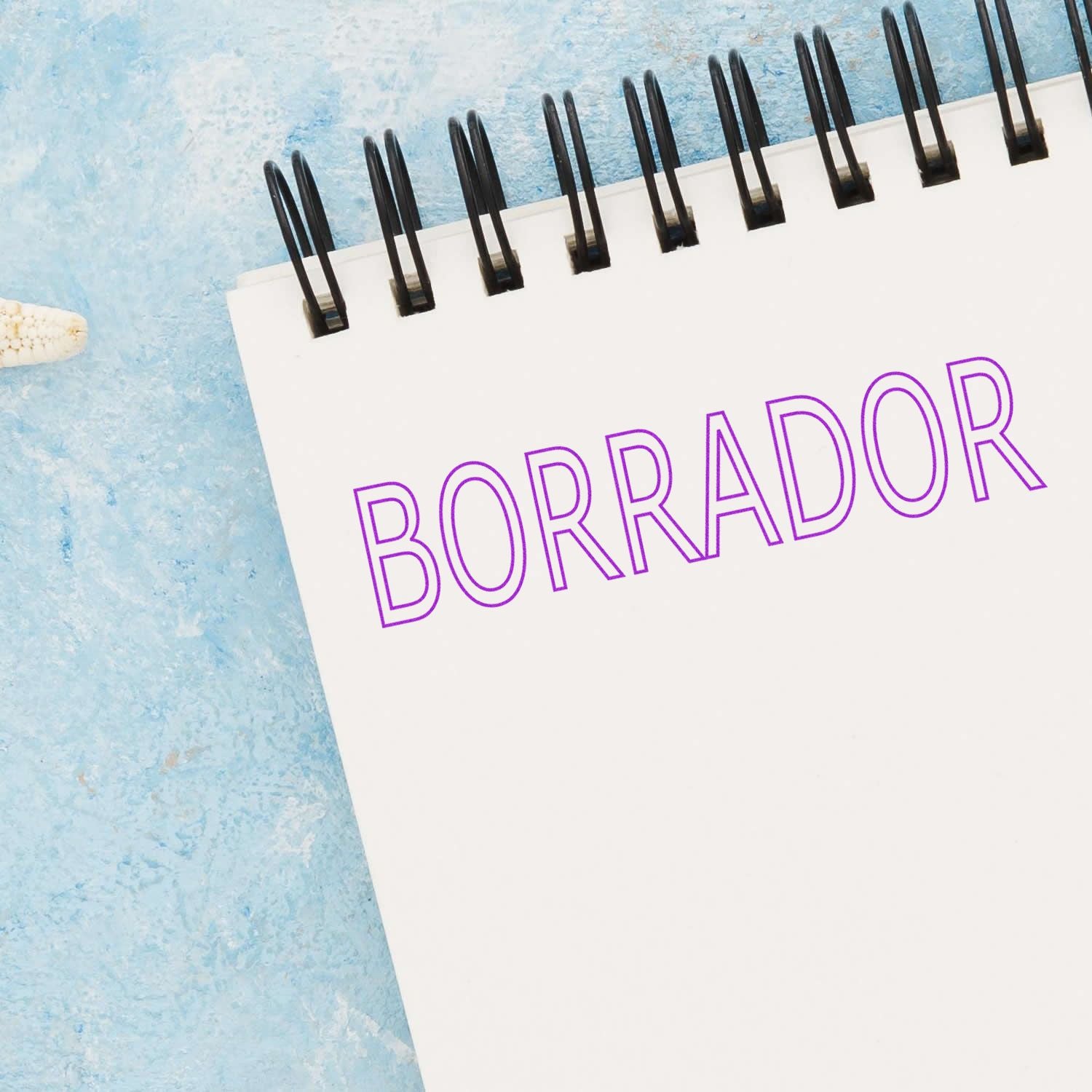 A Large Borrador Rubber Stamp imprinting the word BORRADOR in purple ink on a white spiral notebook against a blue background.